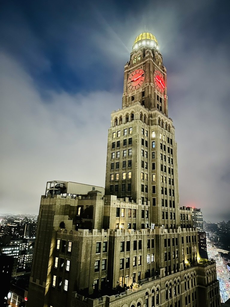 Nighttime view of a tall clock tower building with illuminated clocks showing around 9:02, featuring a golden dome at the top and city lights in the background.