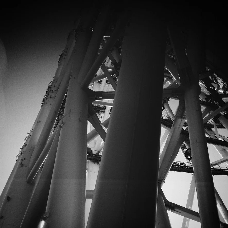 Black and white photo of a massive Ferris wheel structure with large supporting beams and ferris wheel cabins seen from beneath.