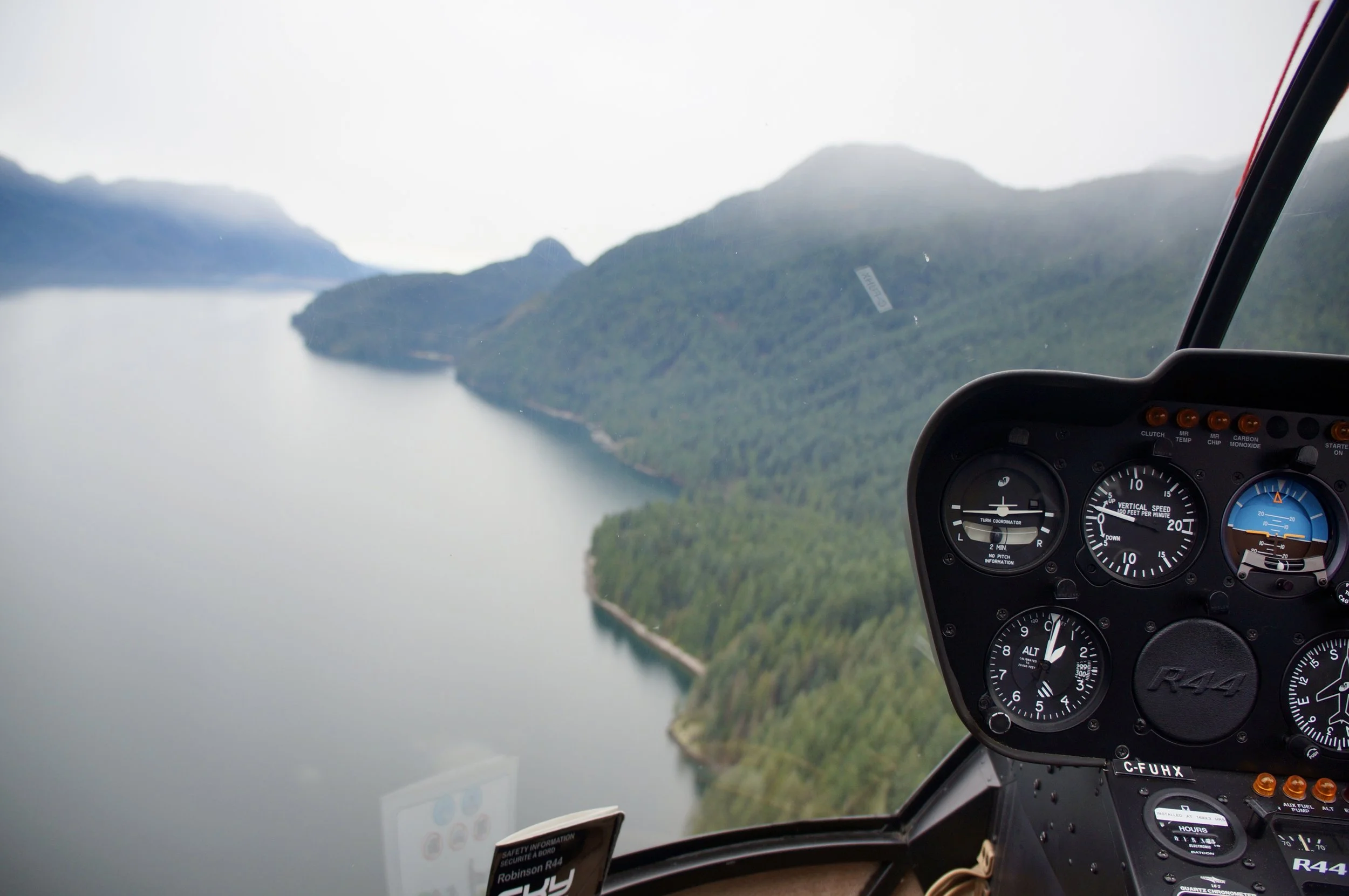 View from inside a helicopter cockpit showing the instrument panel with gauges, with a landscape of mountains and a river below.