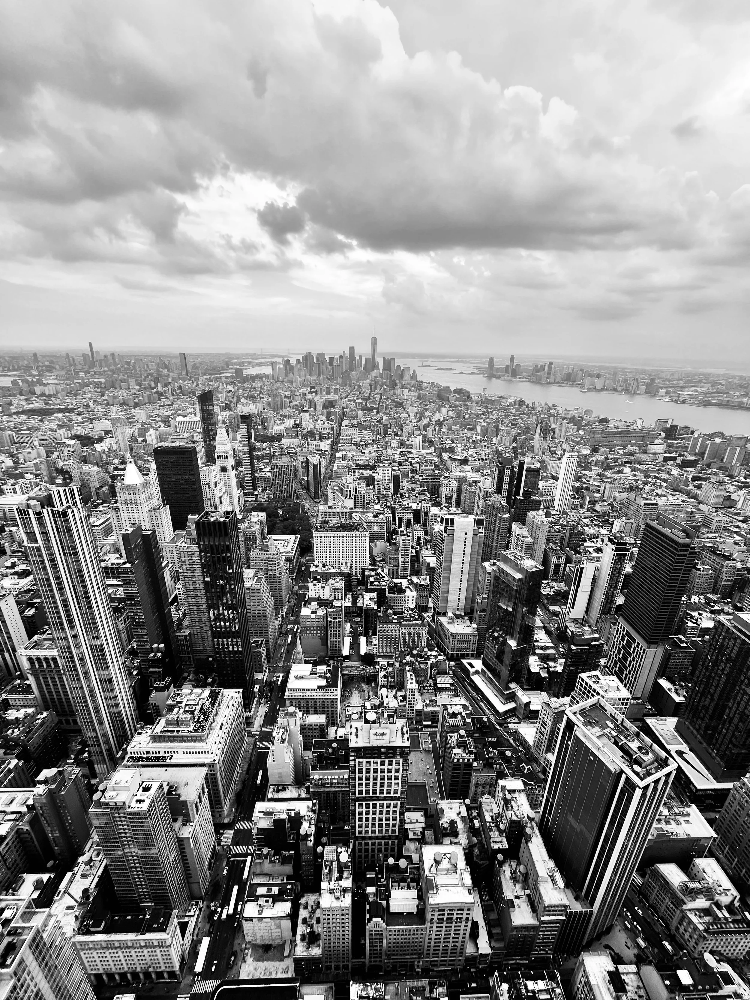 Aerial view of New York City skyline in black and white, showing skyscrapers, a river, and cloudy sky.