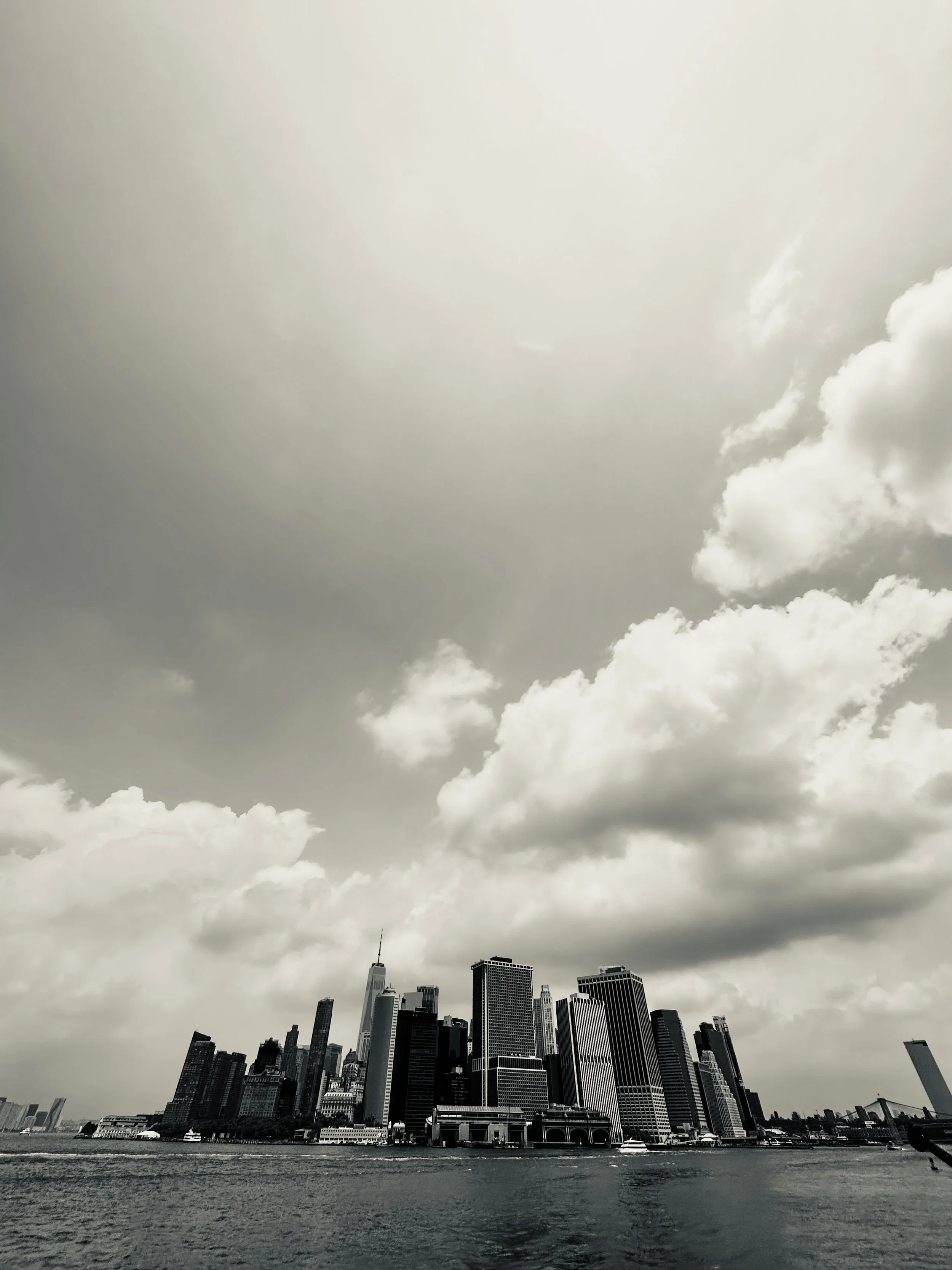 Black and white photo of a city skyline along a river with tall buildings, and a cloudy sky overhead.