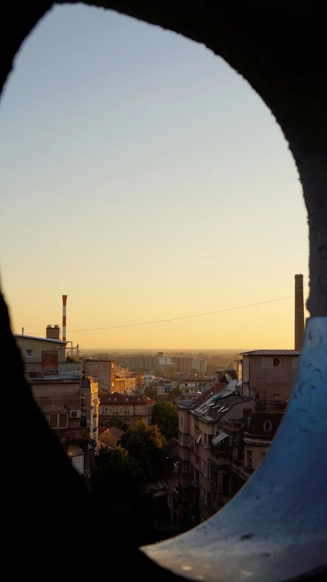 View of Belgrade at sunrise, seen through a circular opening, with buildings and a tall smokestack in the background.