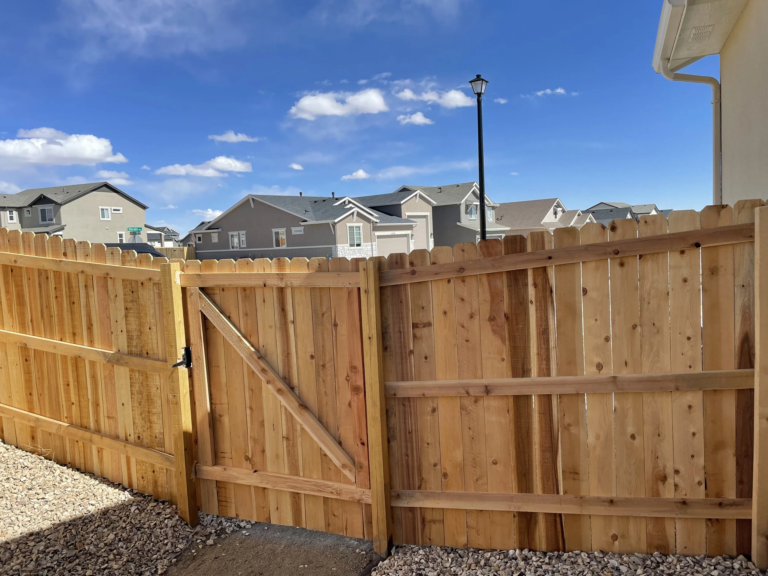 Wooden privacy fence with a gate in a suburban neighborhood, blue sky with scattered clouds, houses in the background, and a gravel ground.