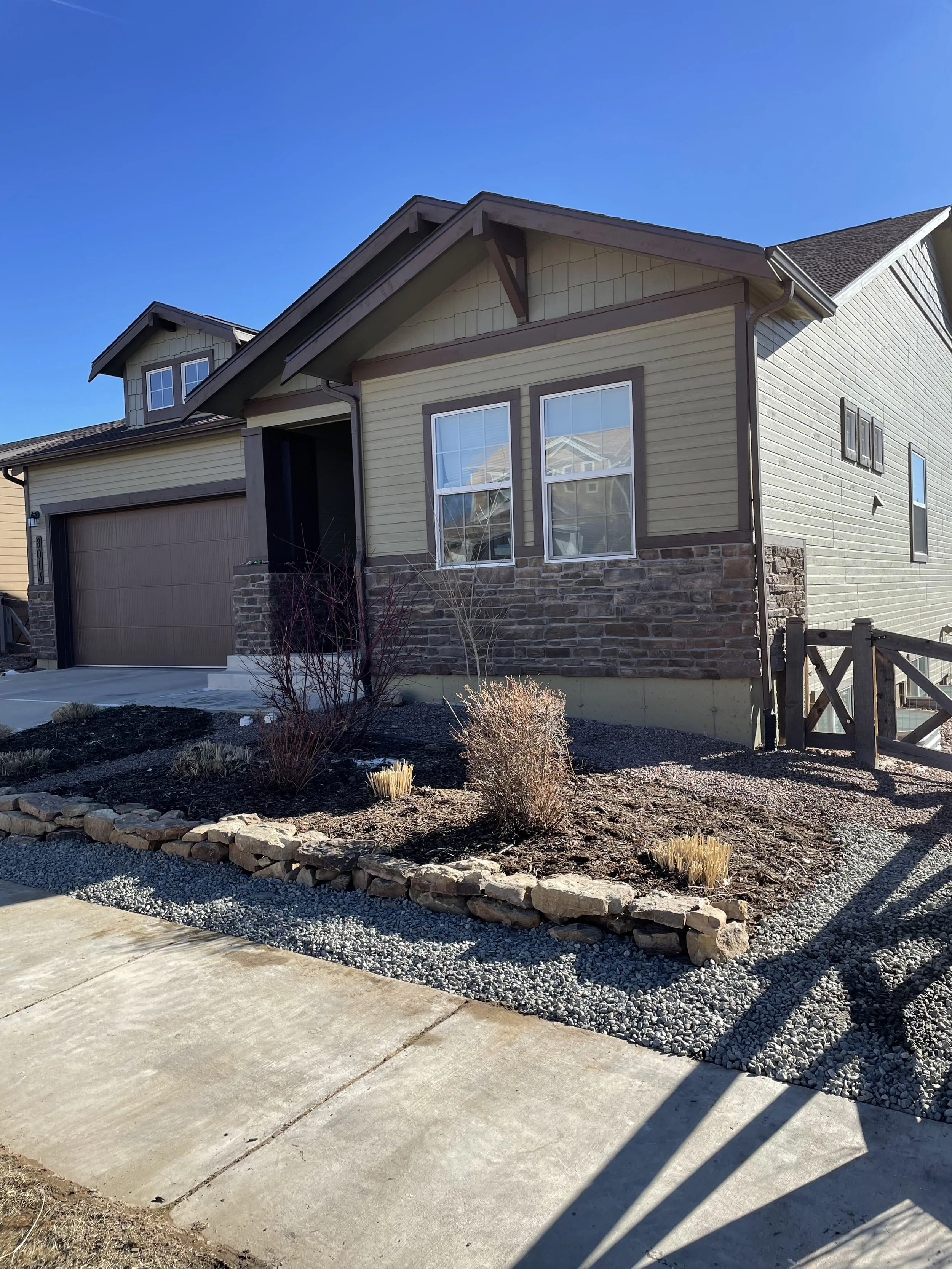 Front yard with small landscaped garden, shrubs, and stone border in front of a beige house with stone accents and a two-car garage. Clear blue sky.