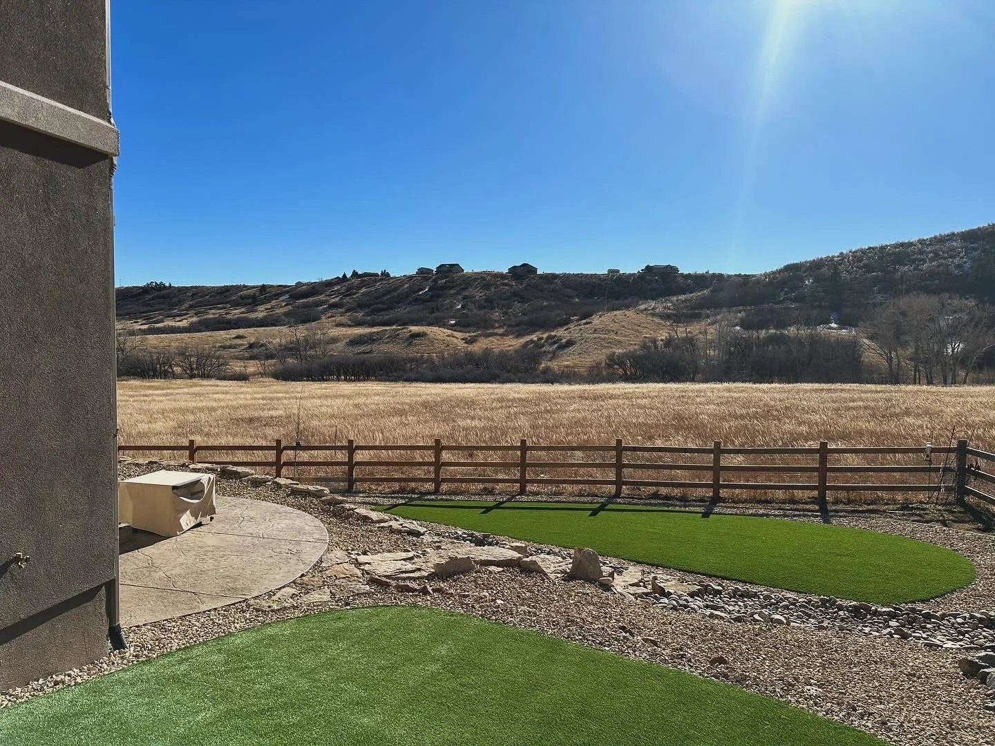 Backyard with artificial turf patches, stone pathway, wooden fence, rural landscape with hills and houses under clear blue sky.