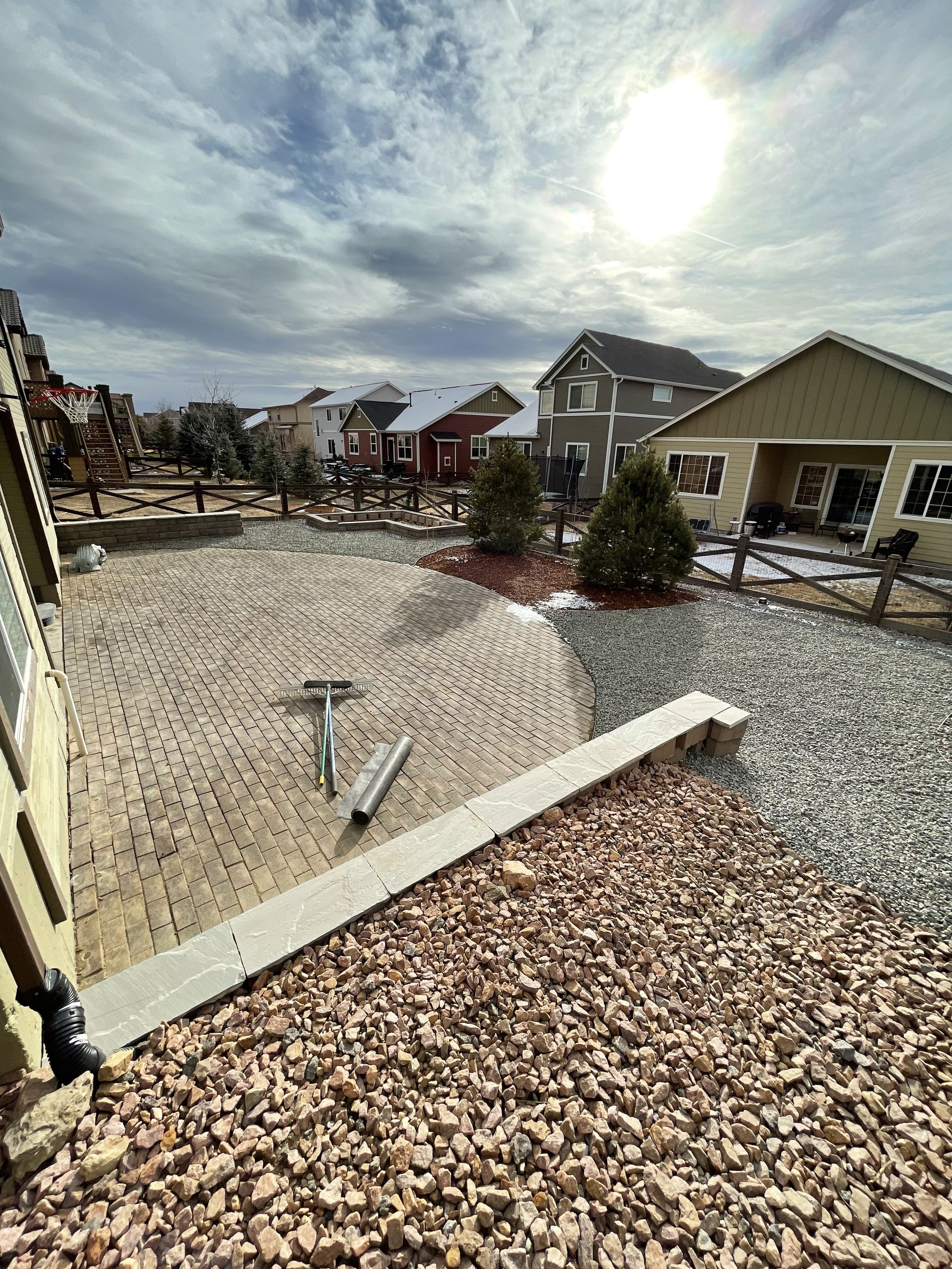 A backyard with a circular brick patio, two small trees, and a gravel area. Today is a partly cloudy day with the sun shining.