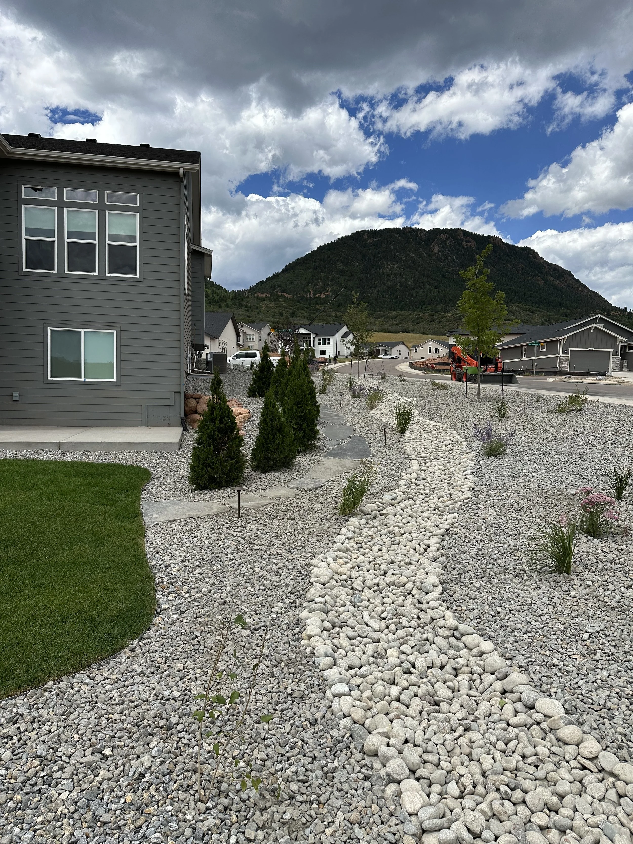 A modern gray house with a rock and gravel front yard, a stone pathway, small bushes, and a lush mountain in the background under partly cloudy skies.
