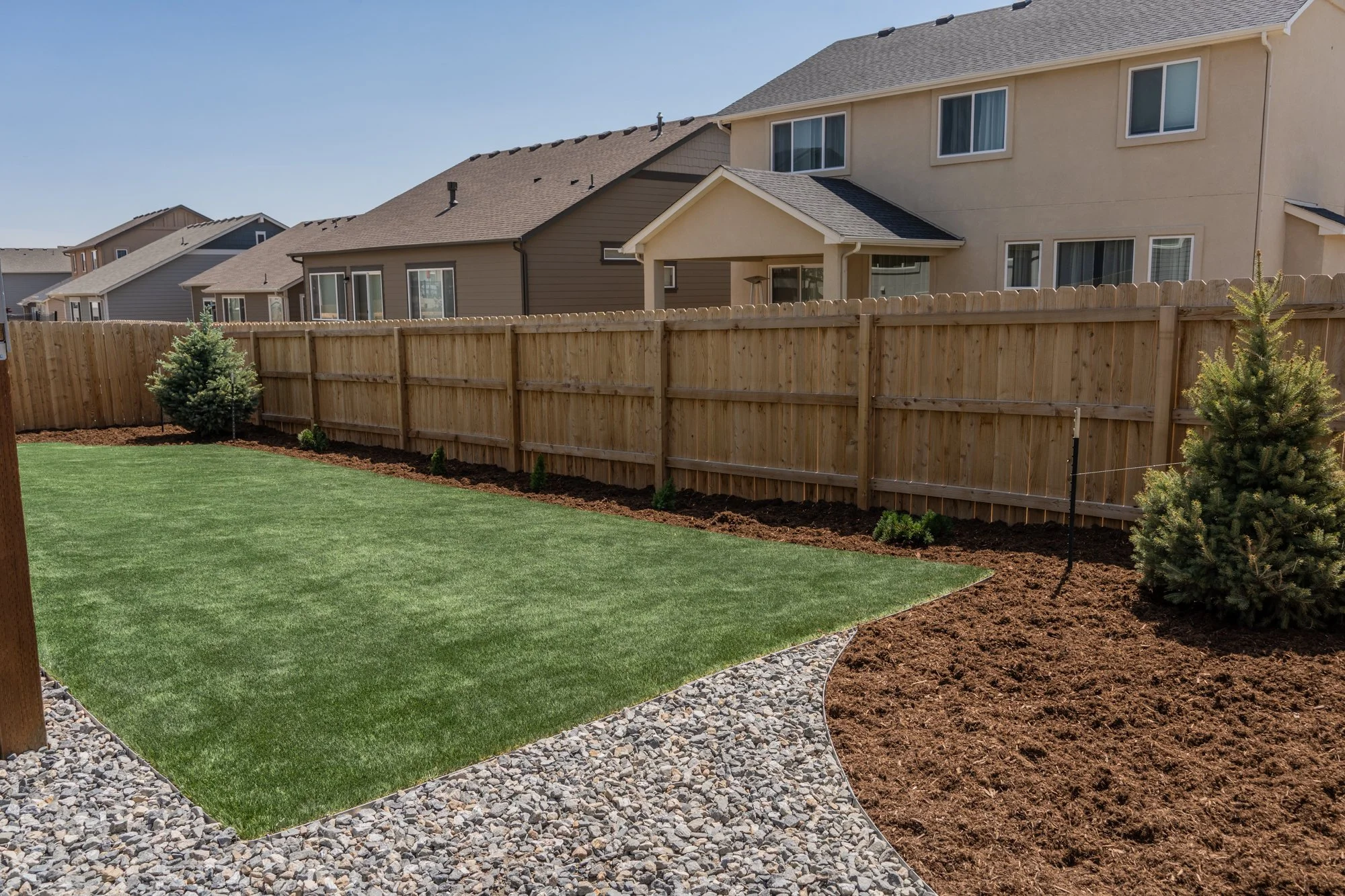 Backyard with artificial grass, landscaped areas, a wood fence, and small trees in a suburban neighborhood.