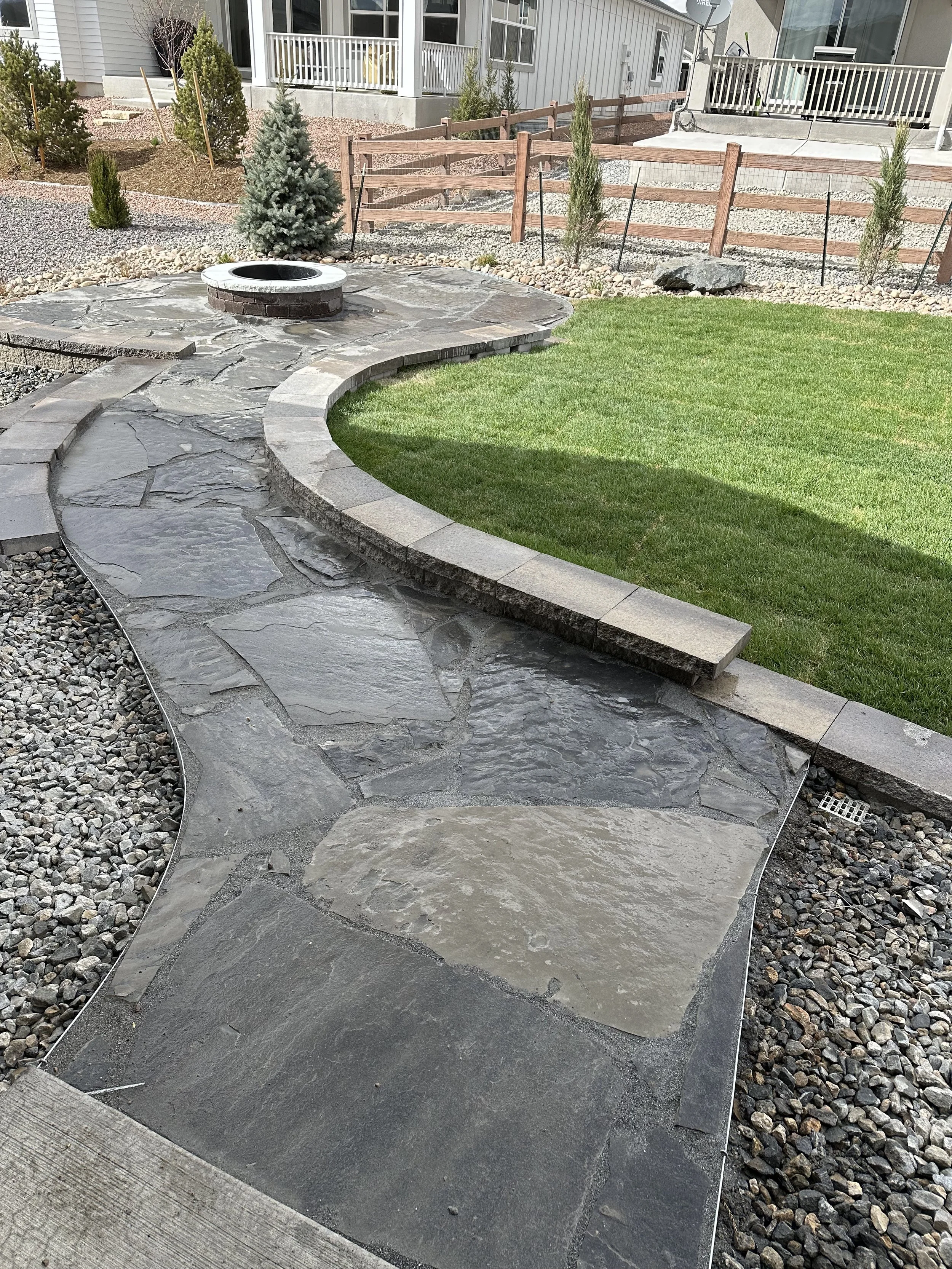 A landscaped backyard with a curved stone pathway, a well-maintained green lawn, small trees, and a circular fire pit. There is a house with a porch in the background.