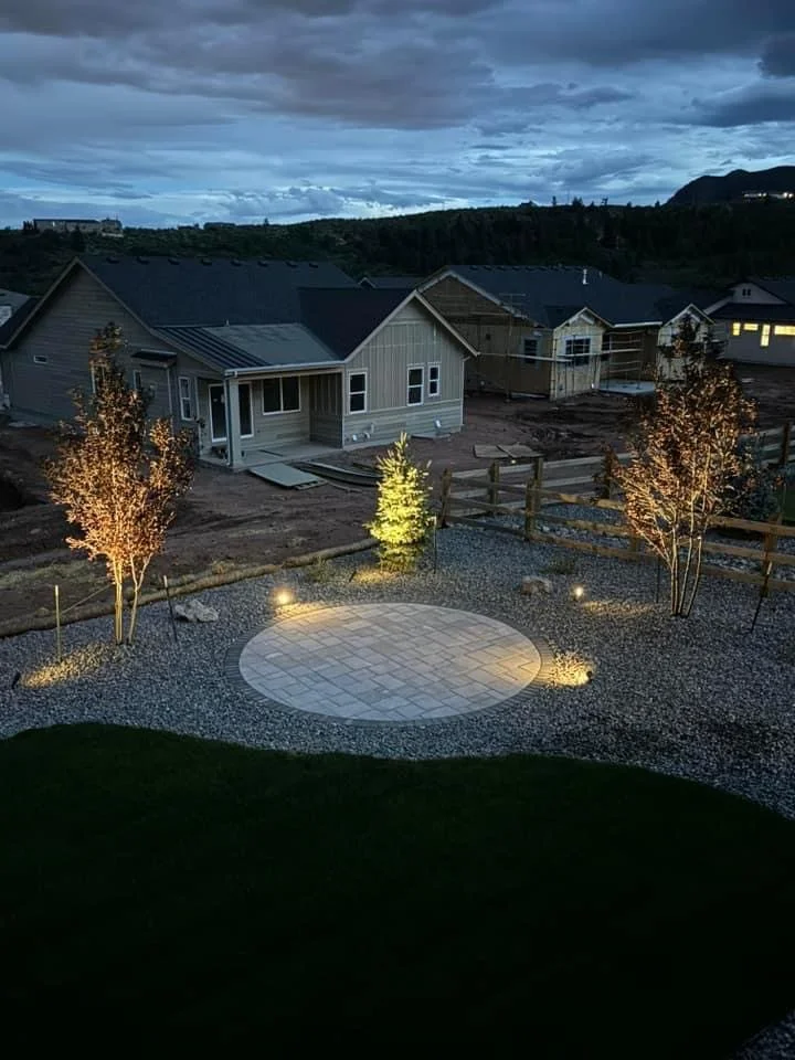 Nighttime view of a backyard with a circular paved area illuminated by outdoor lights, surrounded by small trees and gravel landscaping, with houses and a cloudy sky in the background.