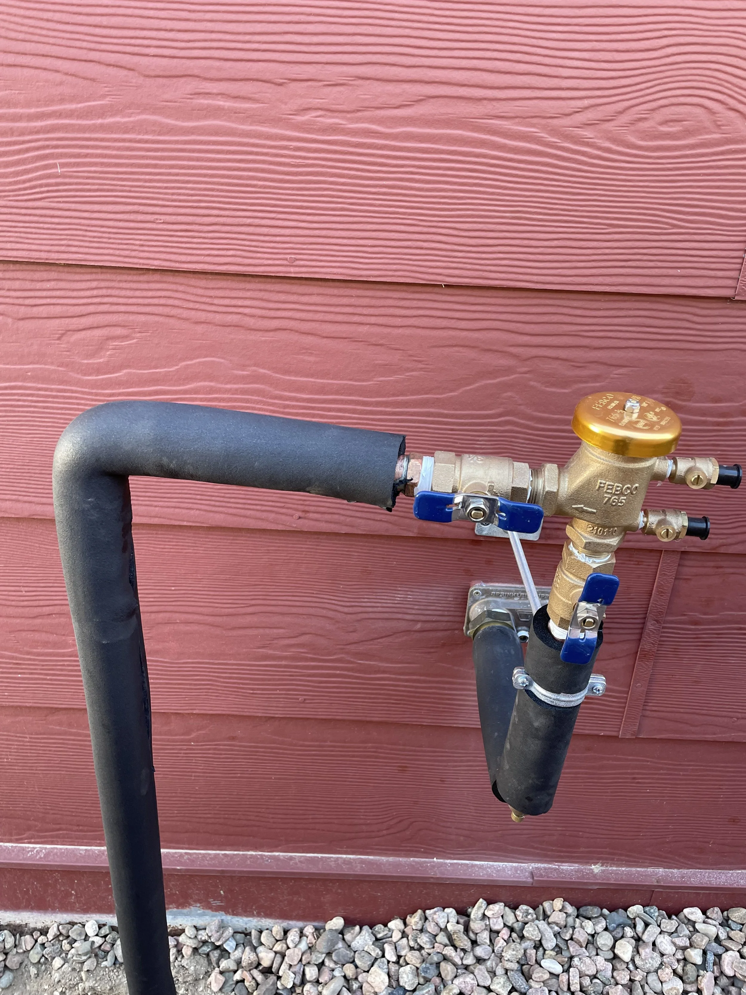 Outdoor plumbing connection with a brass valve, black pipe, and blue handles against a red wooden wall.