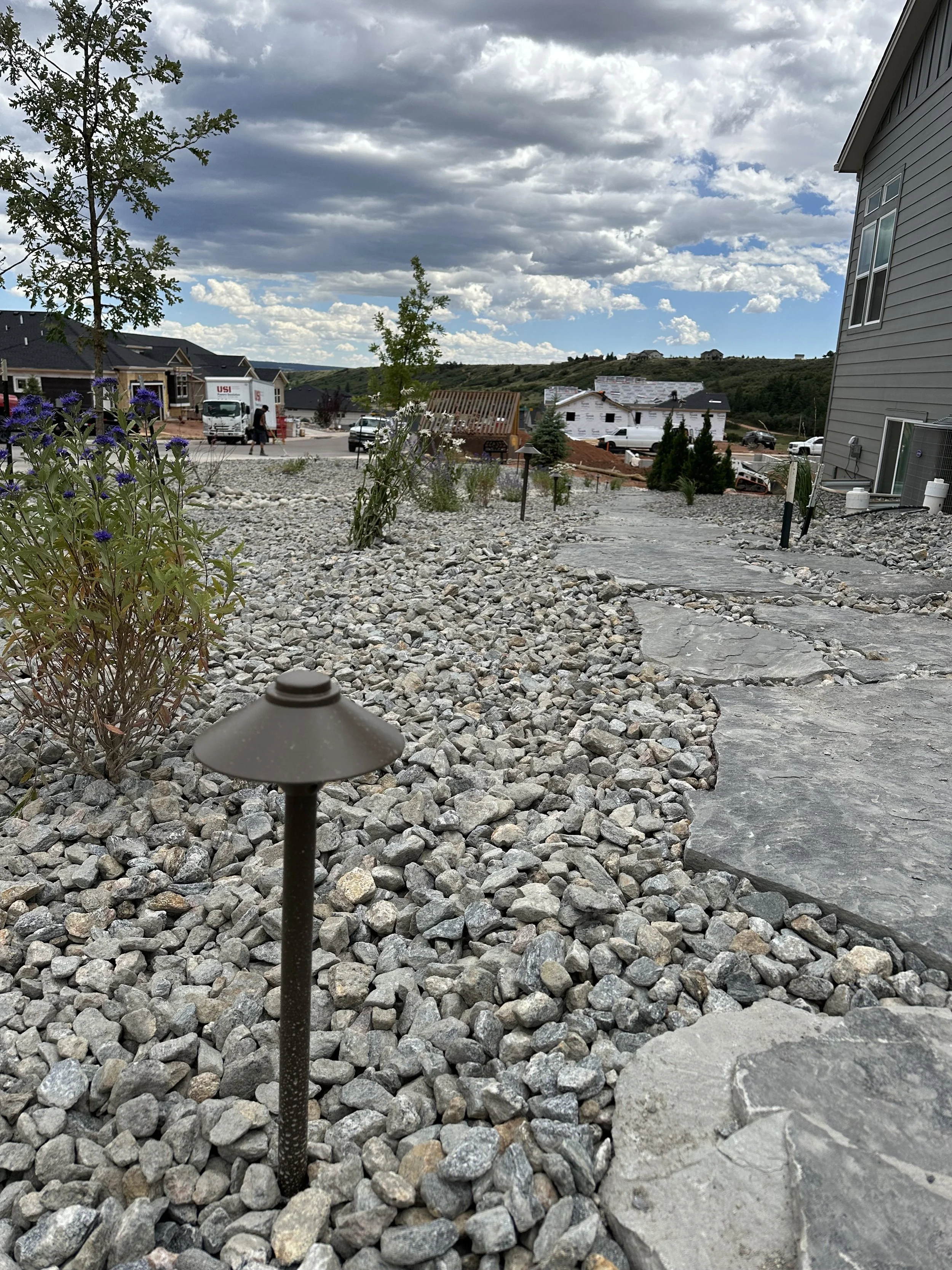 A gravel pathway with small landscaping lights and young trees in a yard under a partly cloudy sky, with new houses and construction in the background.
