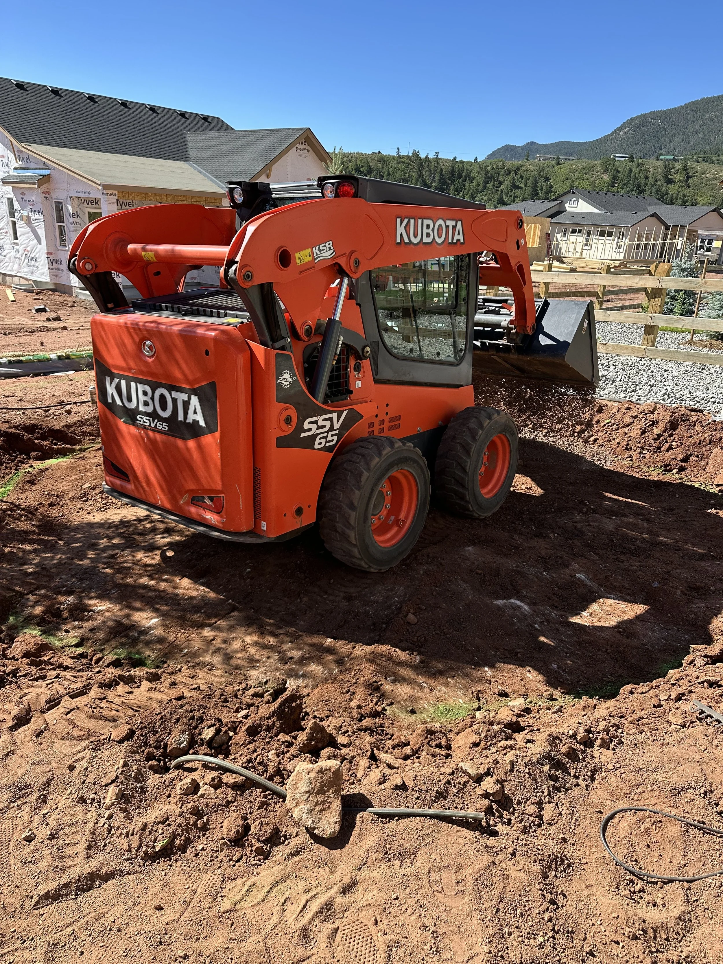A bright orange Kubota skid-steer loader working on a construction site with dirt and gravel around, with residential houses and mountains in the background on a sunny day.