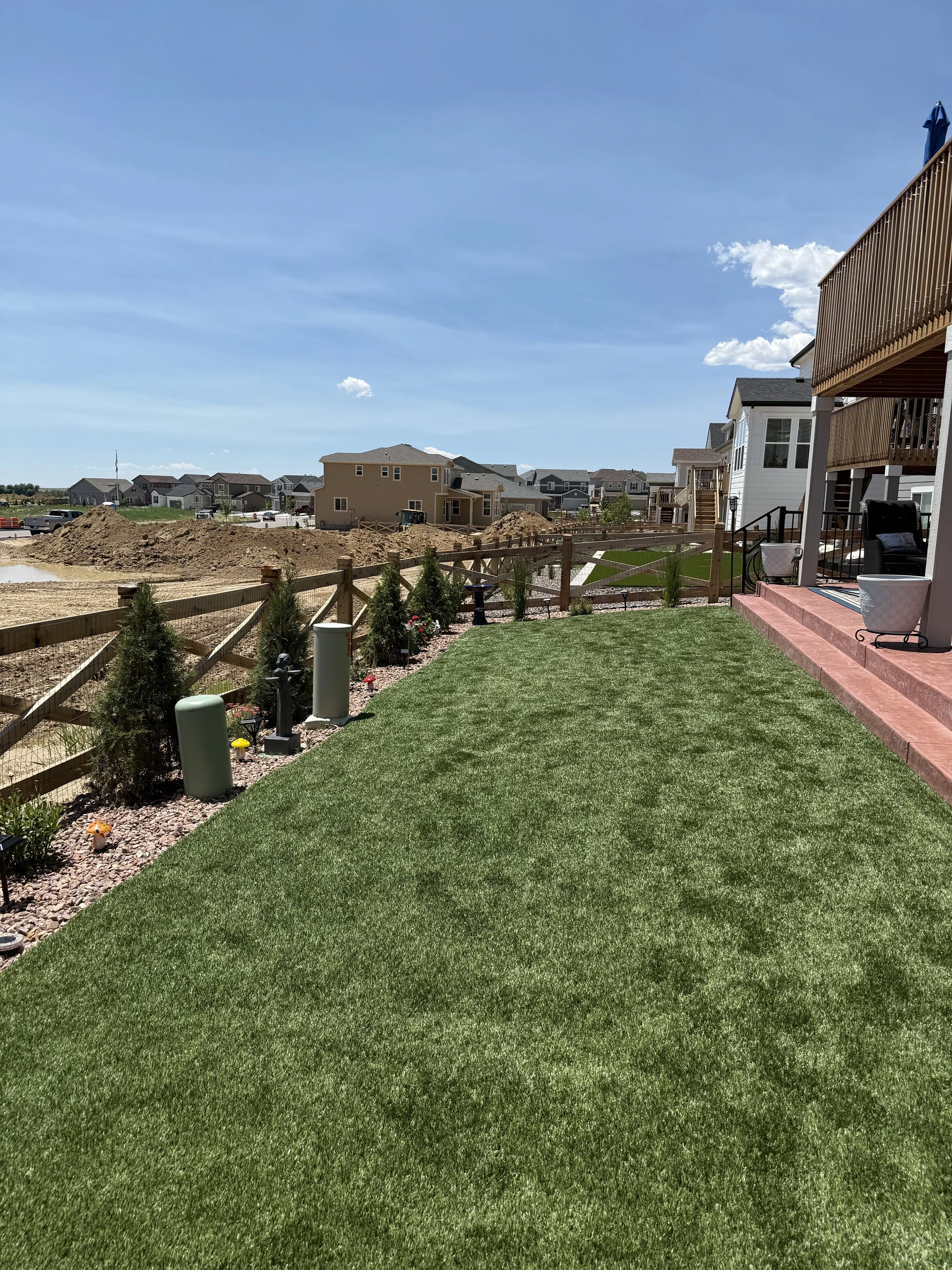 A backyard with a well-maintained green lawn, a wooden fence, small trees and plants along the fence, and a two-story house with a porch and outdoor furniture. In the distance, there are other houses, a construction site with dirt, and a blue sky wit