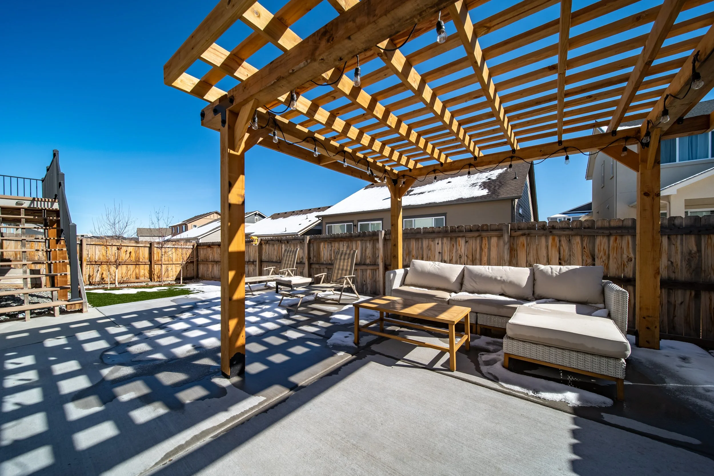 Backyard patio with wooden pergola, outdoor sofa, coffee table, lounge chairs, snow patches, fencing, and neighboring houses under clear blue sky.