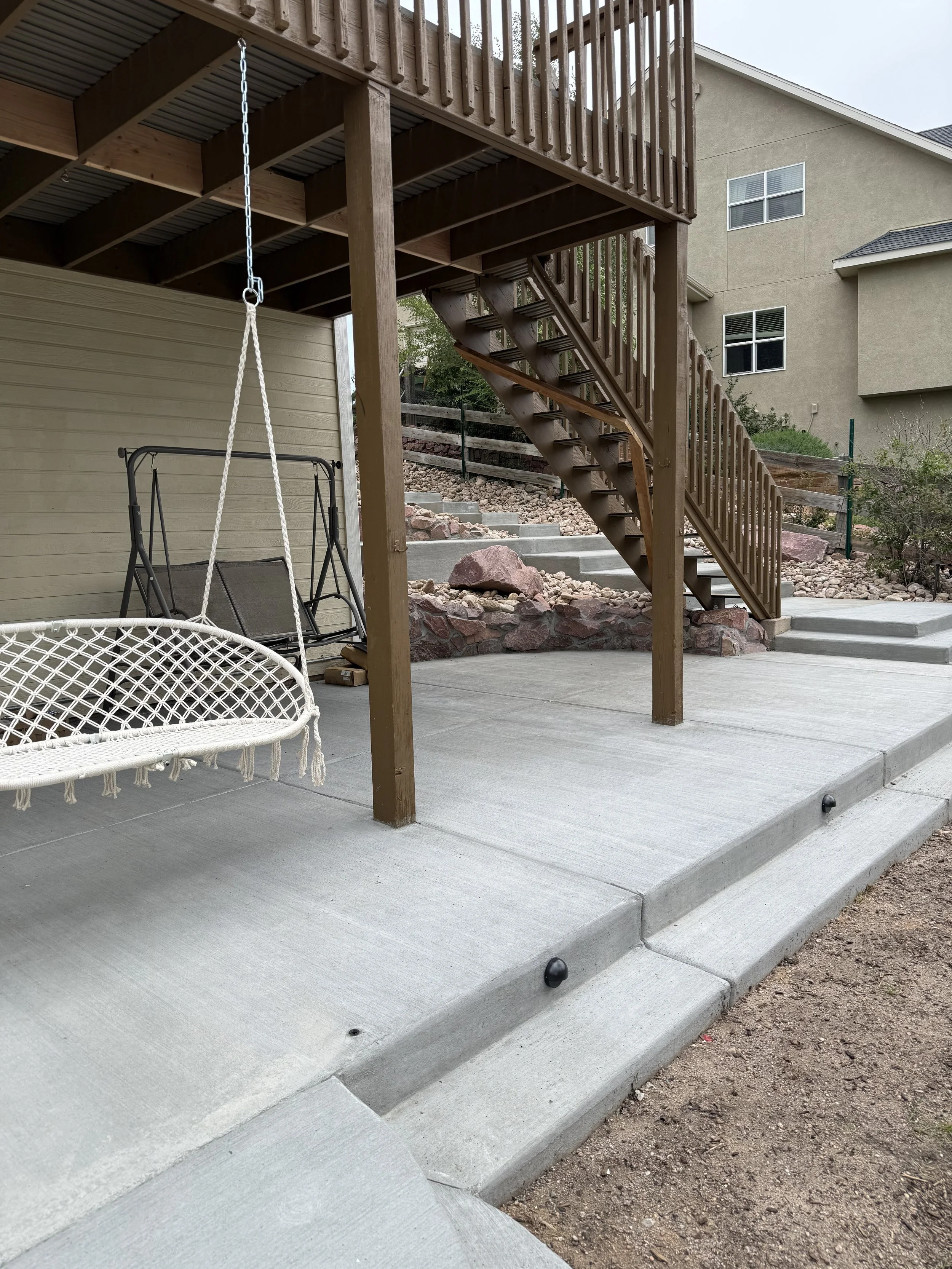 Backyard patio with a concrete surface, a wooden staircase with railing leading to an upper deck, a white hanging hammock swing, a garden swing chair, landscaped rocks, and a house in the background.