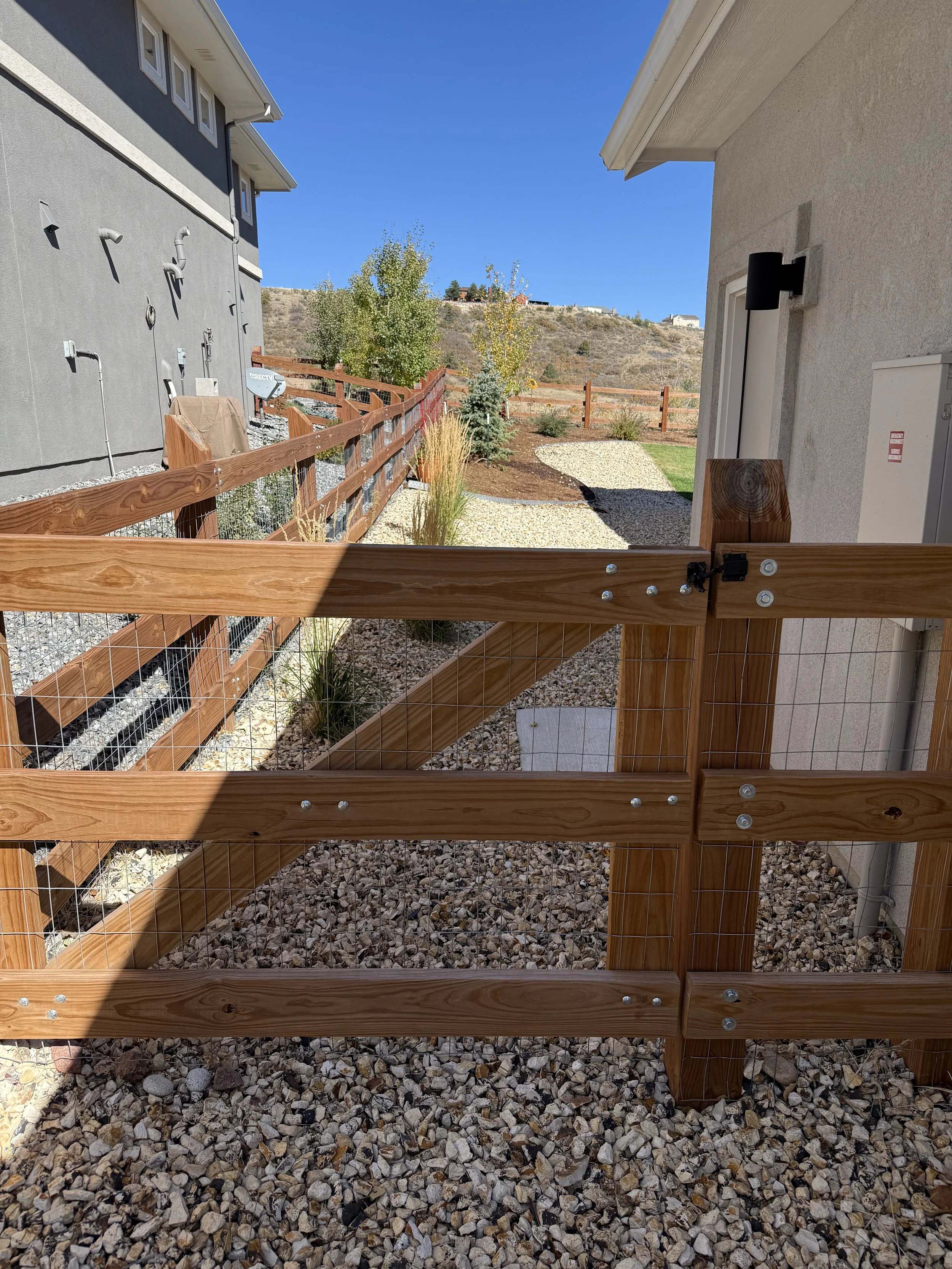 Photo of a backyard with a wooden fence, a gravel pathway, trees, and a hill in the background under a clear blue sky.