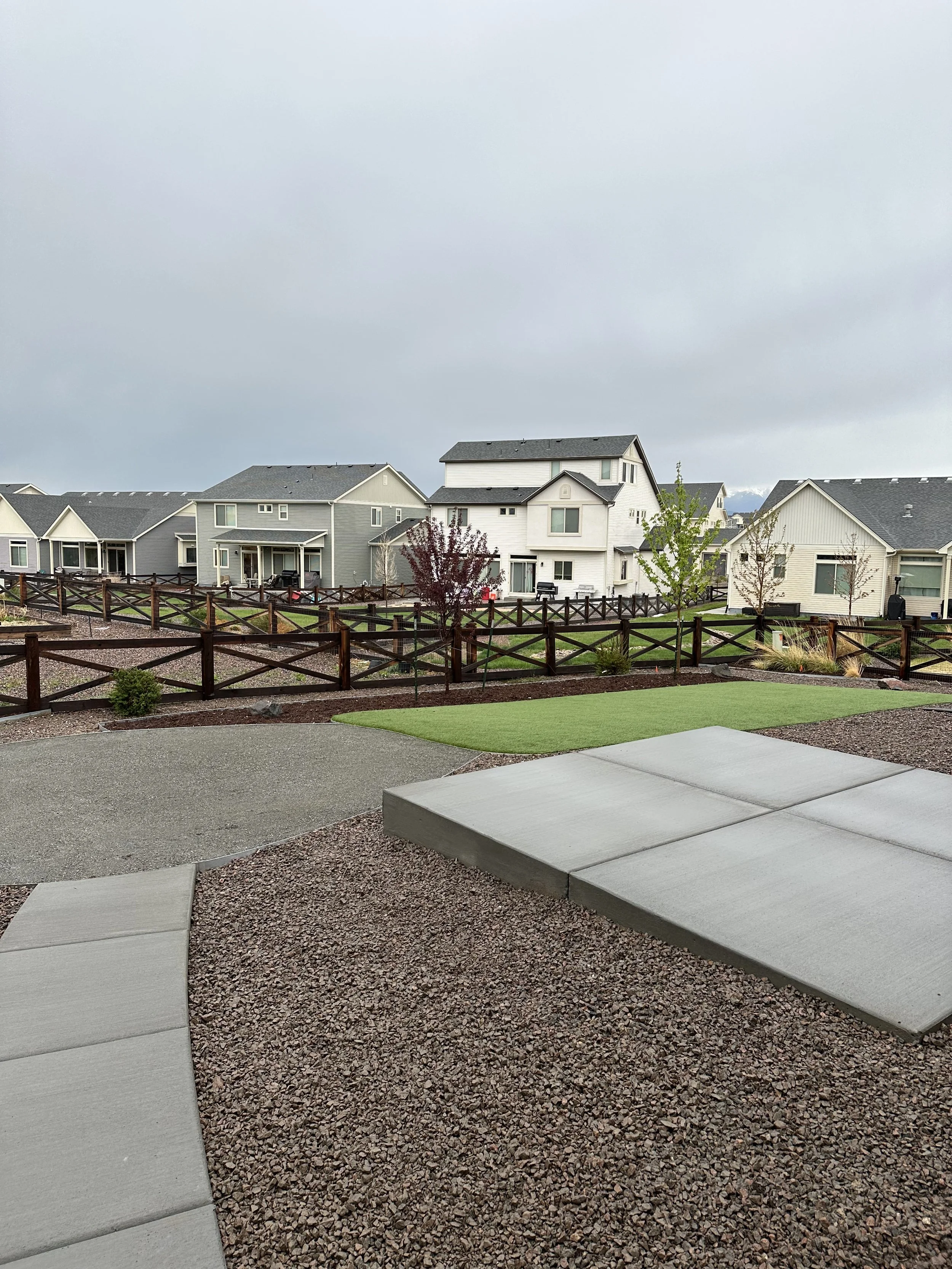 Backyard with concrete patio, artificial grass, and a wooden fence, neighboring houses on a cloudy day.