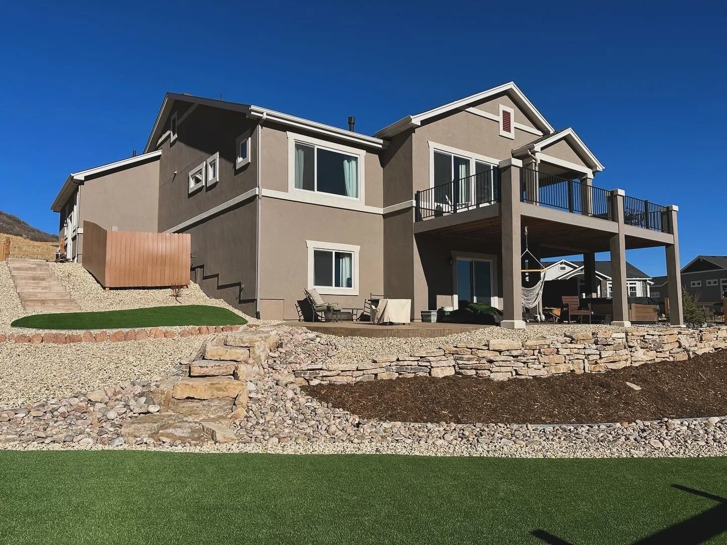 Backyard view of a multi-story beige house with a large deck, sliding glass doors, and outdoor furniture, including a hammock, under a clear blue sky.