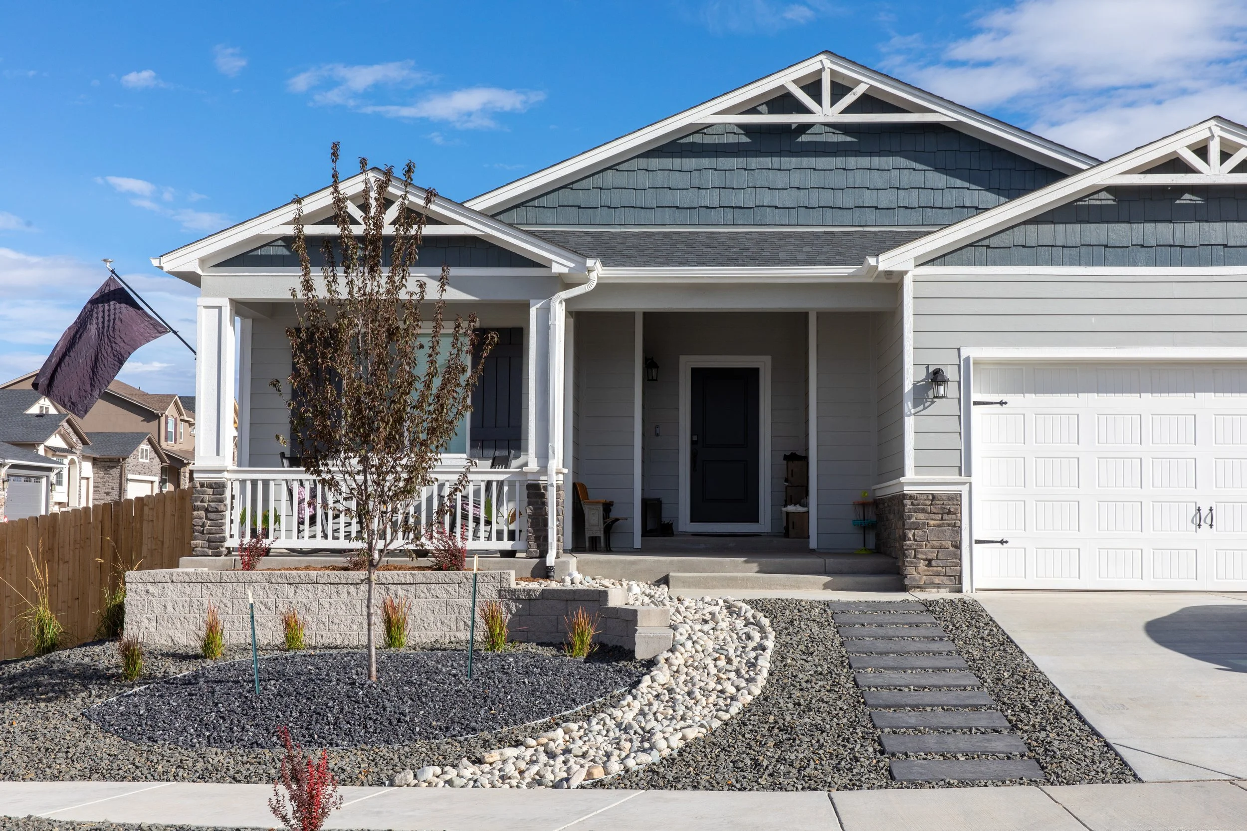 Front view of a modern house with gray siding and a covered porch, landscaped yard with a small tree, decorative rocks, and a walking path, under a partly cloudy sky.