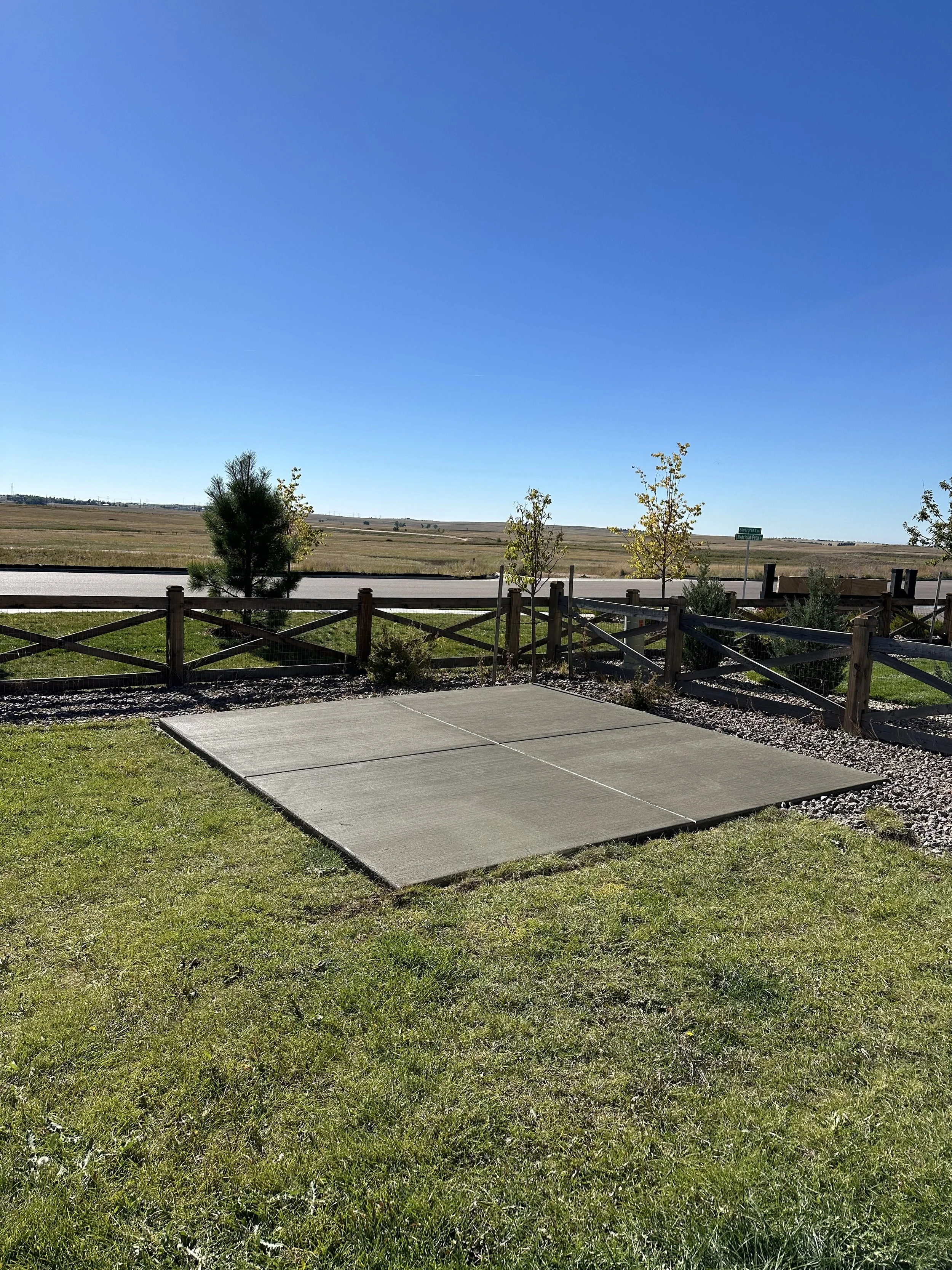Outside concrete patio space with a green grassy area, a wooden fence, and a road with a rural landscape border, under a clear blue sky.