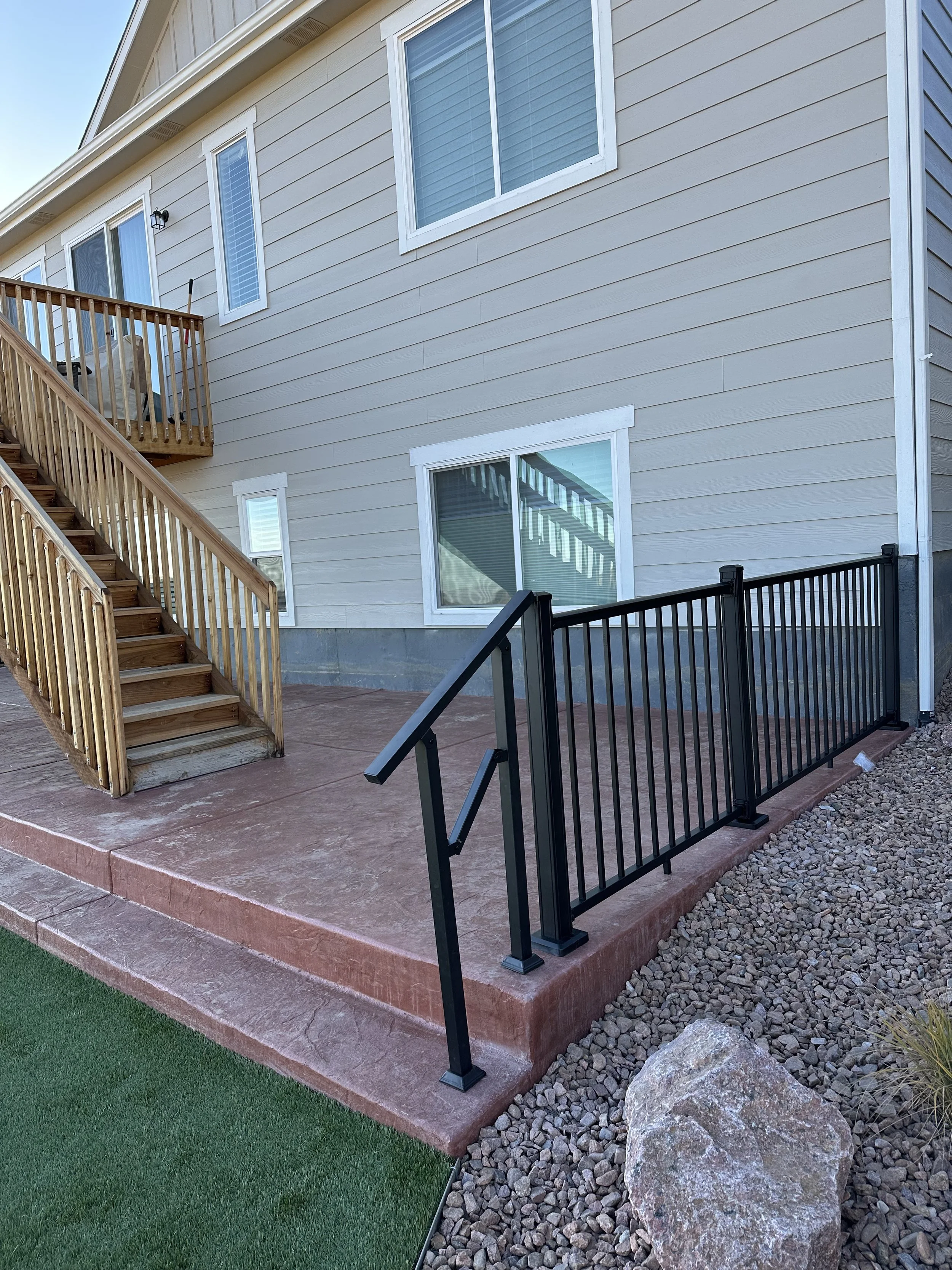 View of a beige house exterior with wooden stairs leading down to a concrete patio, featuring a black metal security fence and a small grassy area with decorative rocks.
