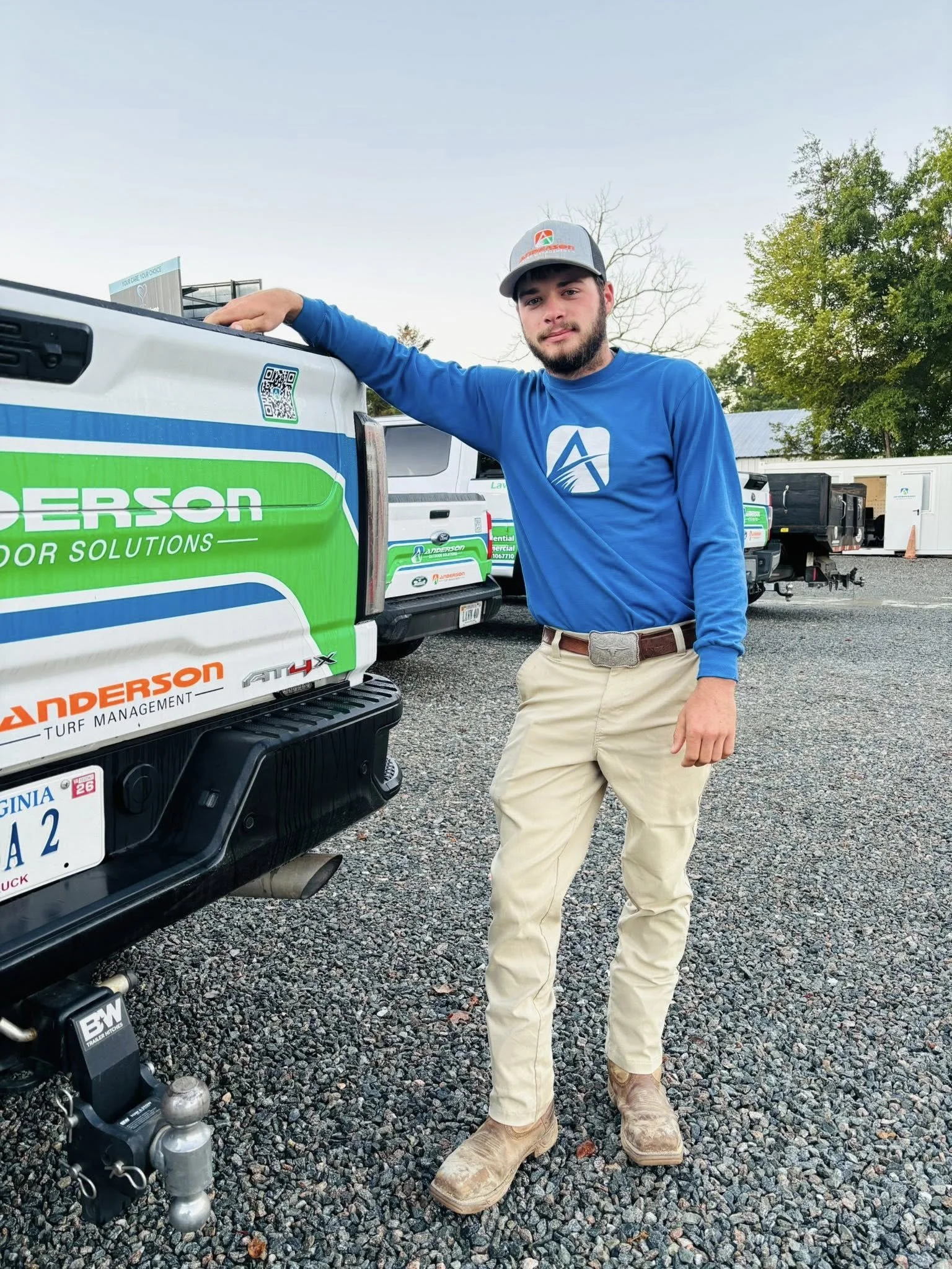 A young man wearing a blue long-sleeve shirt, beige pants, cowboy boots, and a gray baseball cap stands next to a white pickup truck with green and orange lettering. The truck has a Virginia license plate and is parked on gravel. There are several similar trucks and trailers in the background.