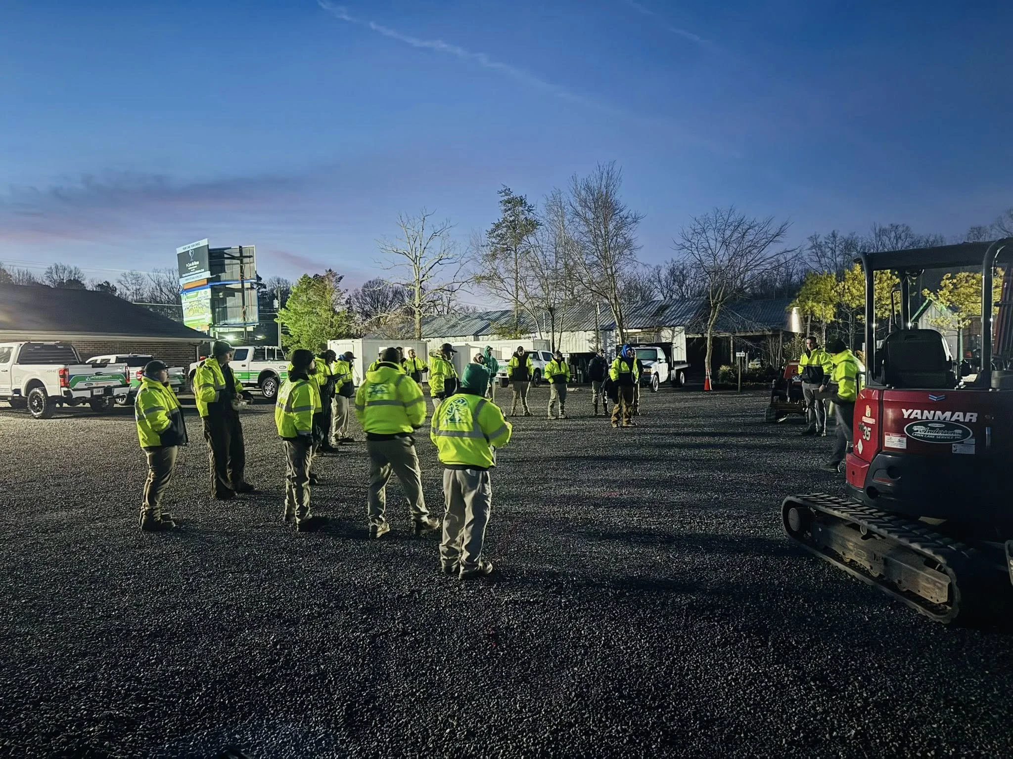 A group of people wearing yellow safety vests standing and listening on a gravel lot with vehicles and trees in the background, with a construction mini excavator on the right.