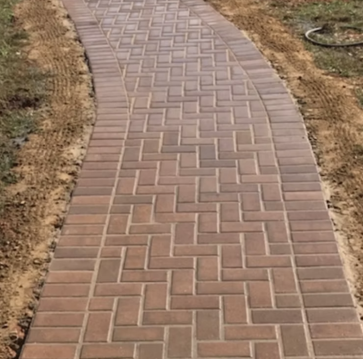 Newly installed brick sidewalk with a curved pattern surrounded by dirt and grass.