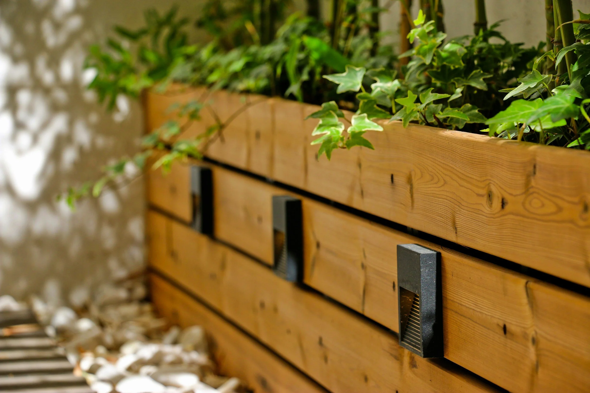 A wooden planter box with green ivy plants, mounted with black metal brackets, inside a landscaped area with rocks and a concrete wall.