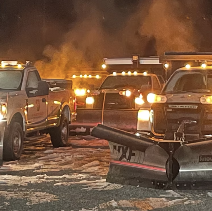 A snow-covered lot with multiple pickup trucks and snow plows, some with lights on, and thick smoke rising in the background.