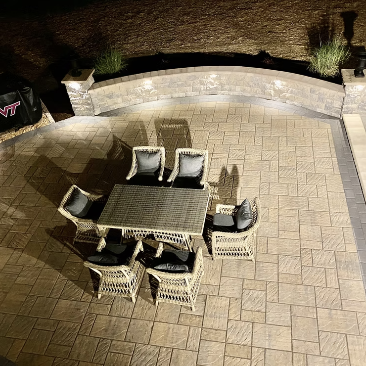 A patio area with a dining table and six wicker chairs with cushions, illuminated at night.