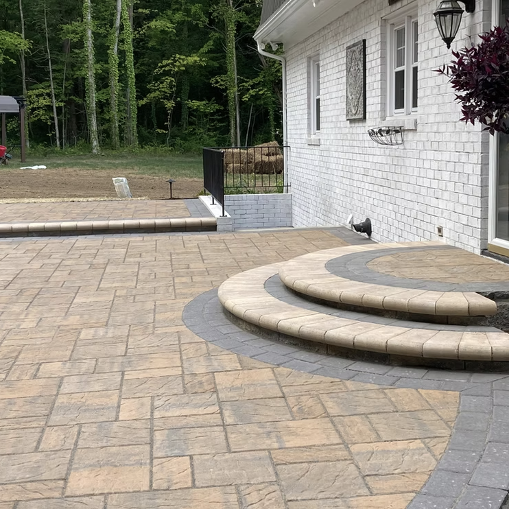 A residential backyard patio with finished stone and paver steps leading to a green yard and wooded area. The patio features a geometric pattern with brown and gray stones, a white brick house with window and wall-mounted decor, and a black metal rai