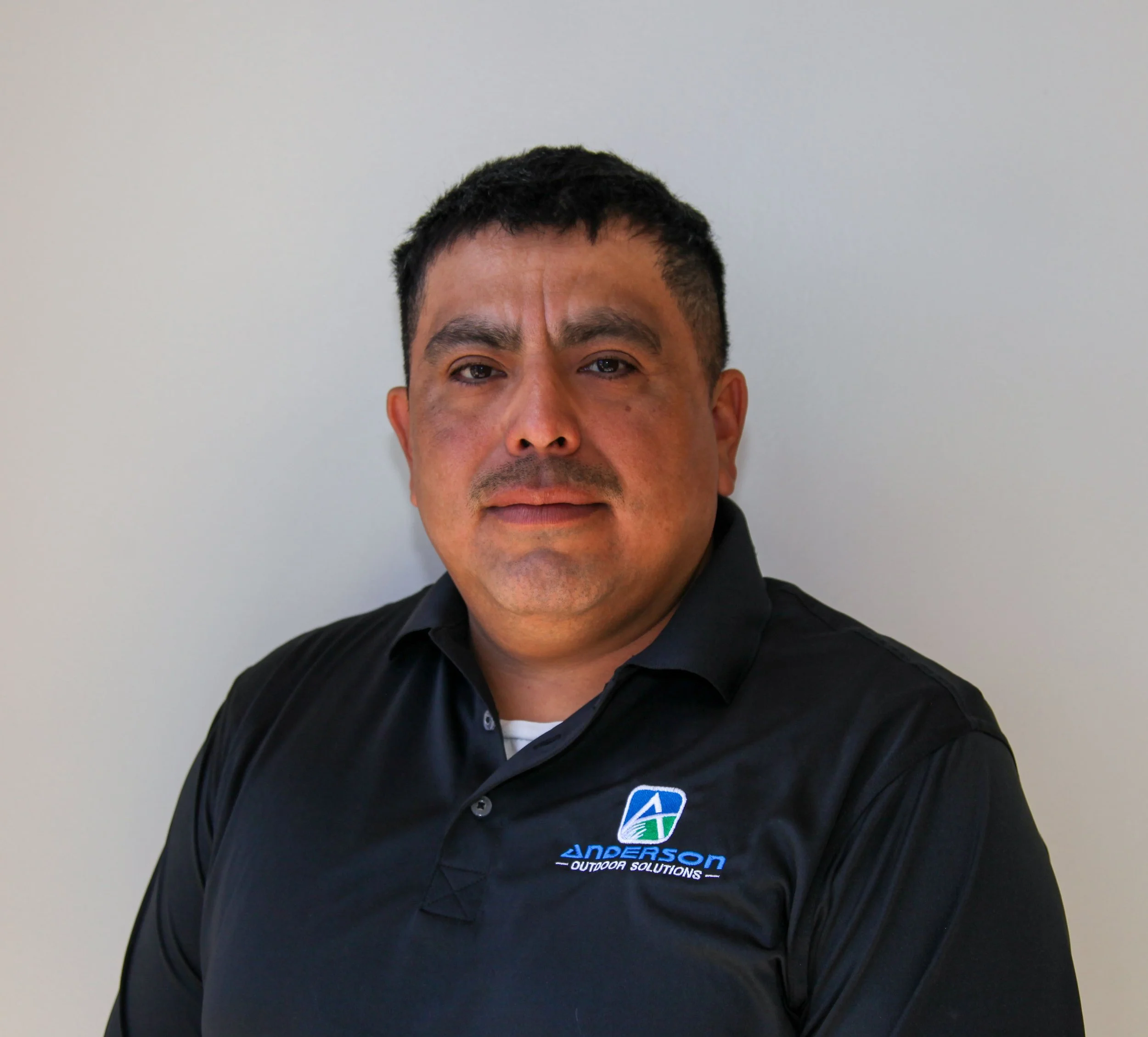 A man with short black hair wearing a black collared shirt with a logo reading 'Anderson Outdoor Solutions' standing against a plain white wall.
