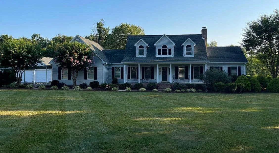 A large house with a front porch, surrounded by well-manicured lawn and bushes, with trees in the background on a sunny day.