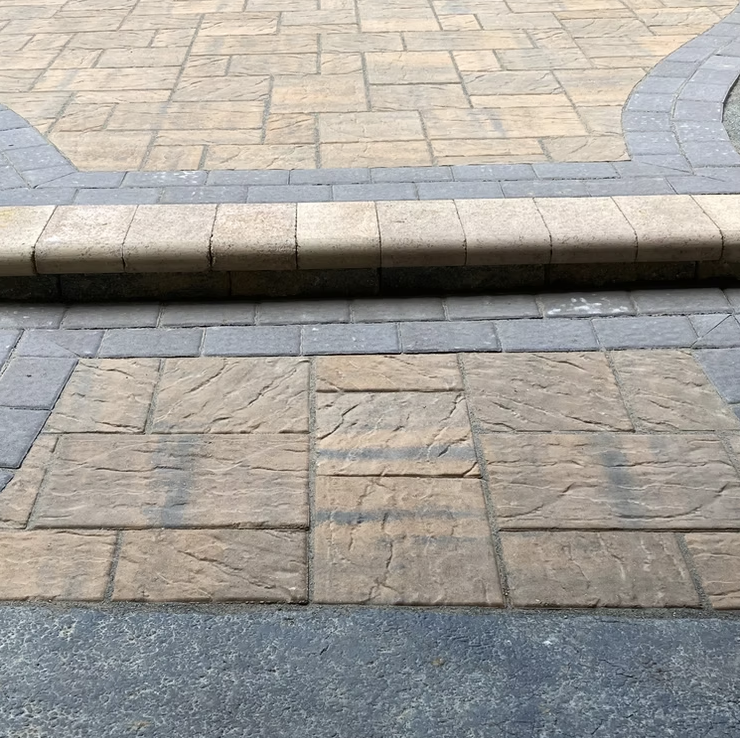Close-up of a paved sidewalk with a small curb or step, featuring tan brick and gray stone tiles.