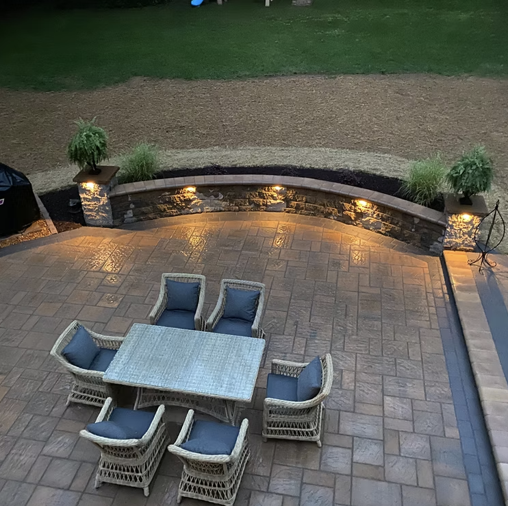 A patio outdoor dining area with a square table and six chairs with blue cushions, surrounded by a brick paving, with small trees and garden beds in the background, illuminated by warm lights.
