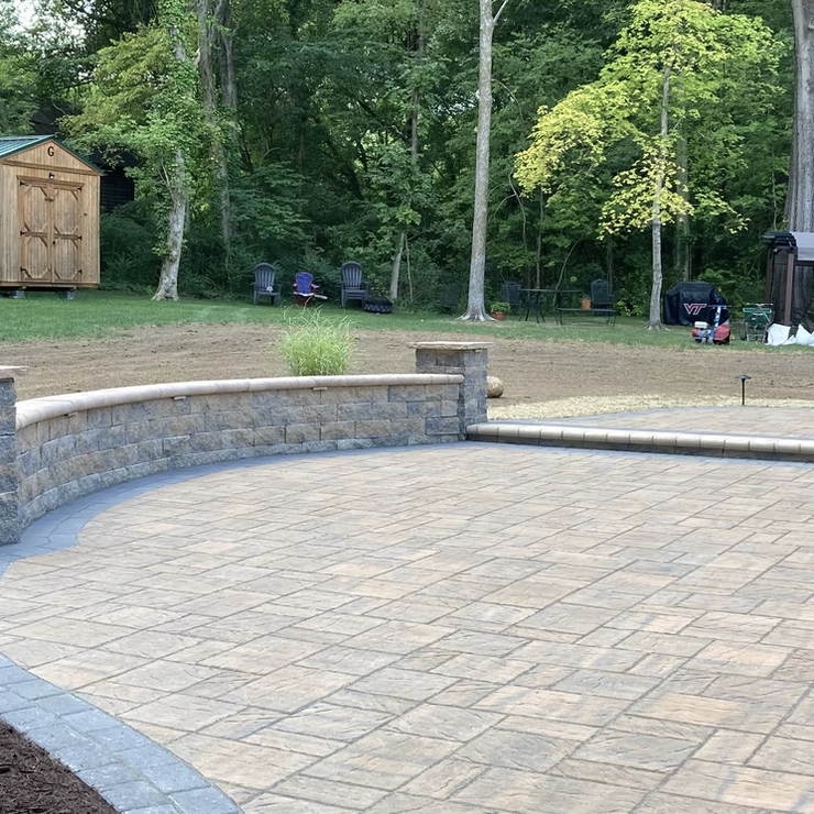 Backyard with newly constructed stone patio, retaining wall, and garden bed, surrounded by lawn and trees.