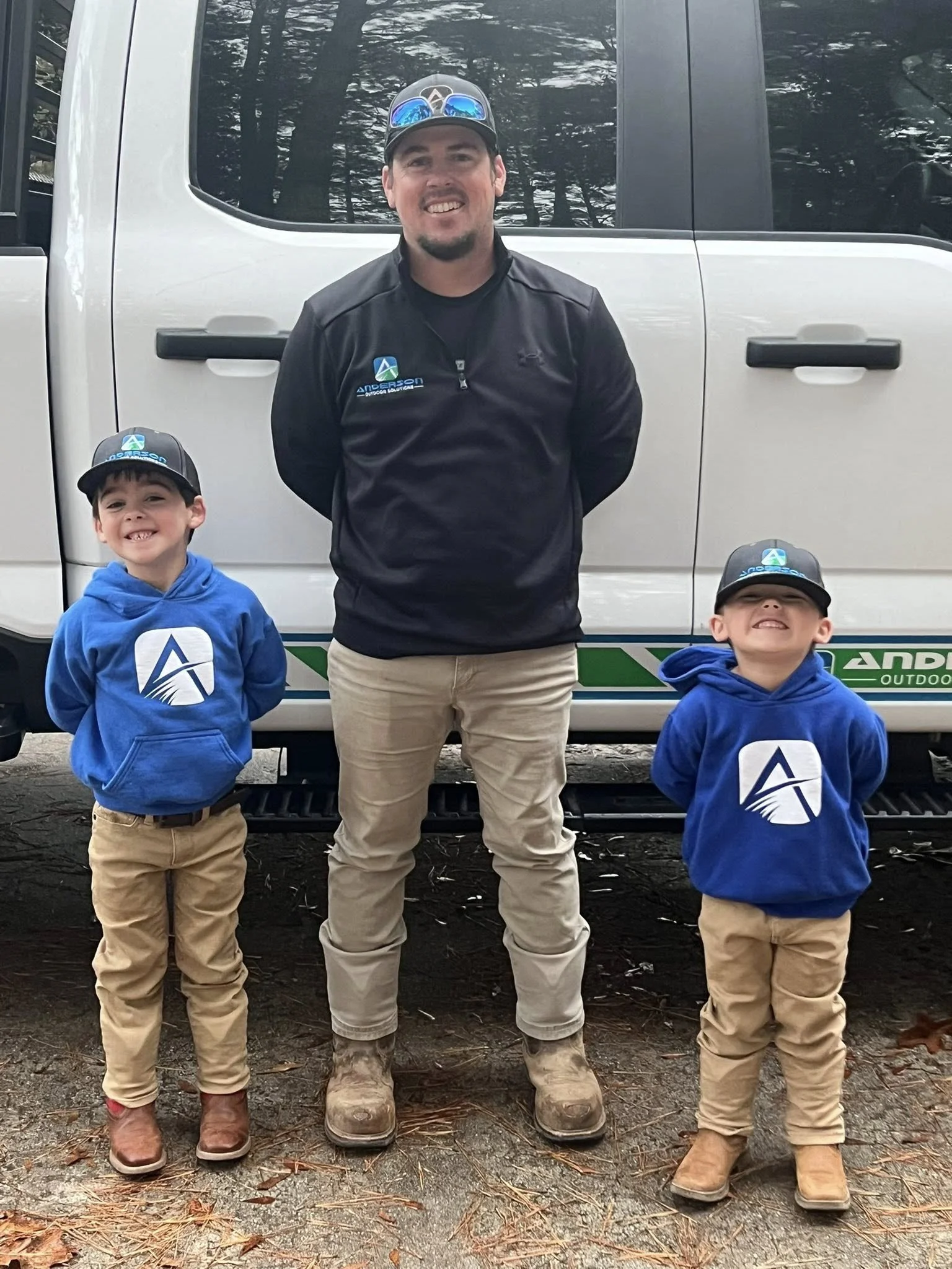 A man with two young boys standing in front of a white truck outdoors during daytime. The man is smiling, wearing a black jacket and beige pants, and the boys are wearing blue hoodies with a logo, beige pants, and hats, also smiling.