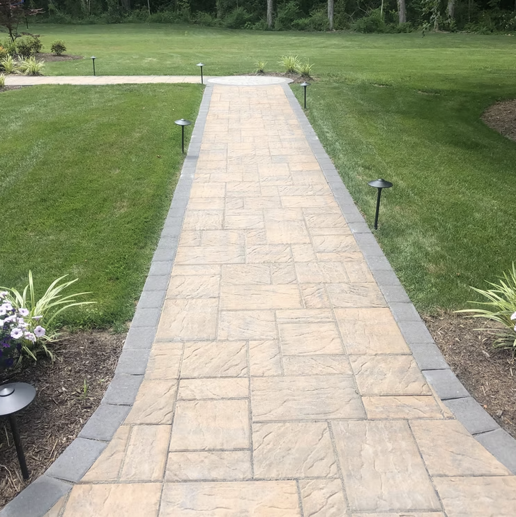 Stone pathway bordered by gray bricks, surrounded by green lawn and garden beds with flowers and plants, outdoor landscape with trees in background.