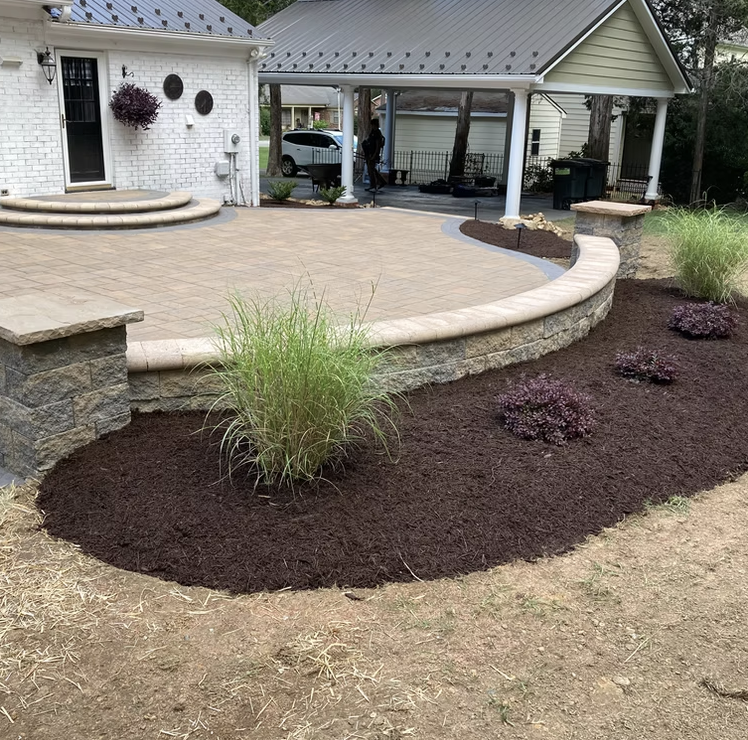 Newly landscaped backyard with a curved stone retaining wall, planted grass and purple shrubs, and a patio with stairs, situated next to a white house with a porch, a car, and a person in the background.
