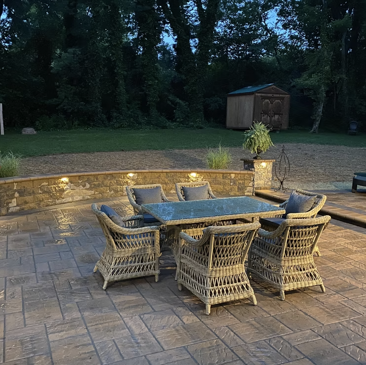 Outdoor patio with wicker dining table and six chairs, illuminated by small lights on a low stone wall, with a grassy yard, trees, a shed, and garden decorations in the background during dusk.