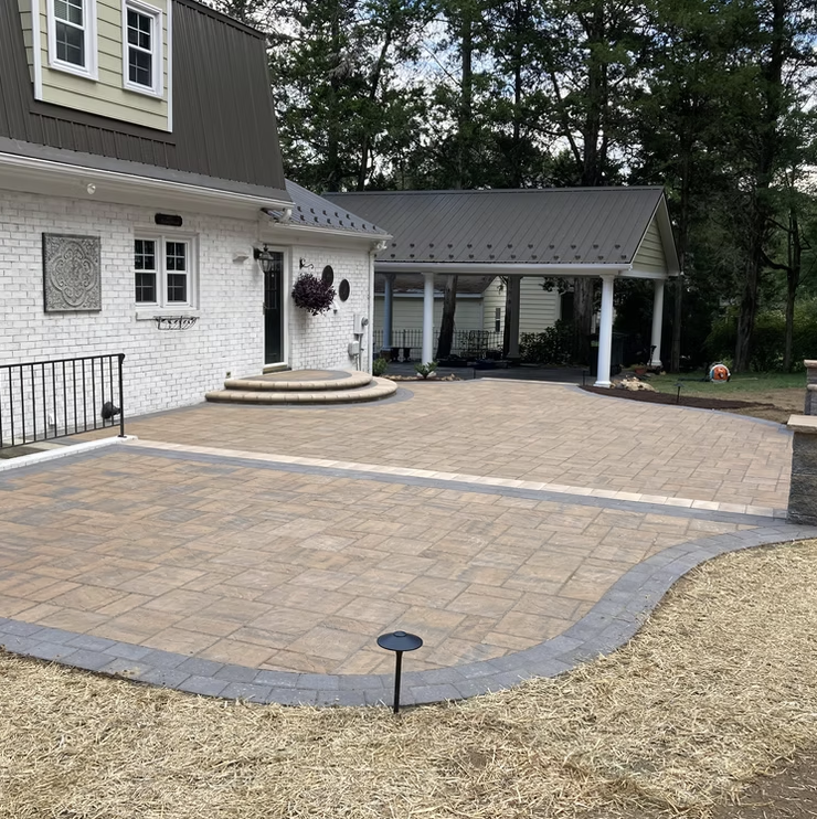 Newly paved backyard patio with brick and stone border, attached to a house with white brick exterior, featuring a garden and trees in the background.