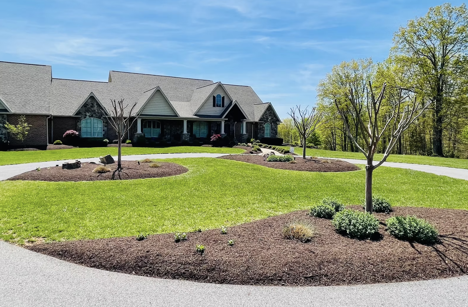 A large house with a peaked roof, stone and brick exterior, front porch, and paved driveway. The front yard has a circular lawn with new trees, small shrubs, and mulch beds under the trees, with a background of trees and a blue sky.