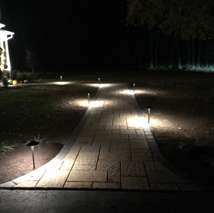 Nighttime view of a paved walkway illuminated by small garden lights along both sides, curving through a yard with trees in the background.