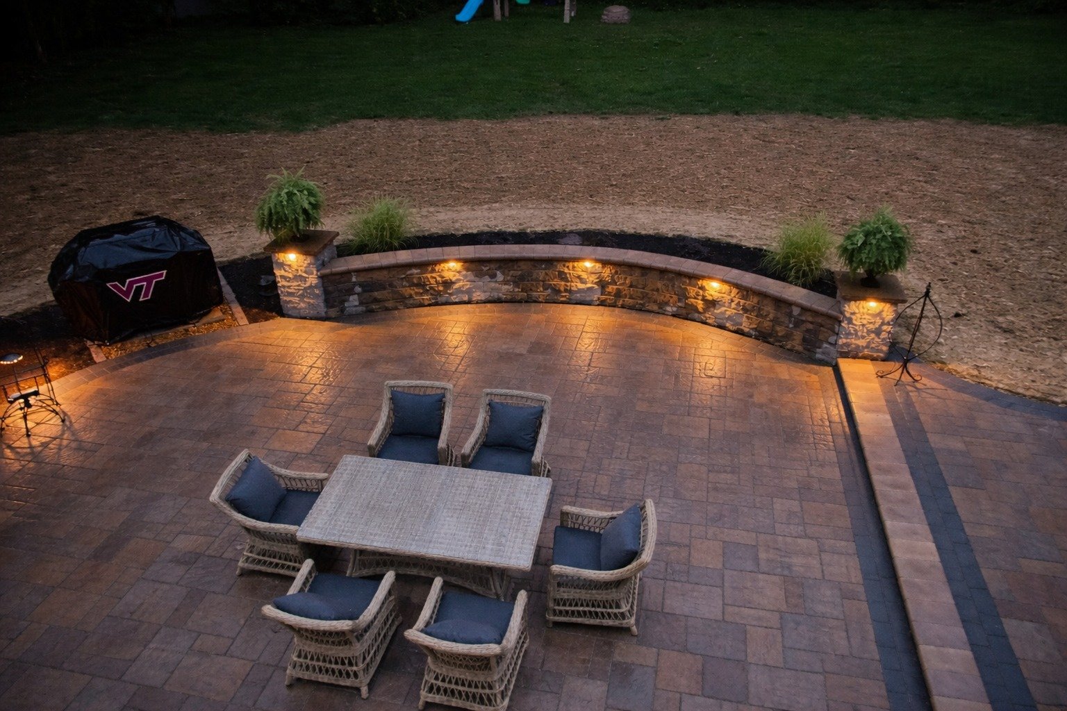 An outdoor patio with a dining table and six chairs, illuminated by built-in landscape lights. The patio is bordered by a curved stone wall with plants, and a grassy yard with a children's playset in the background.