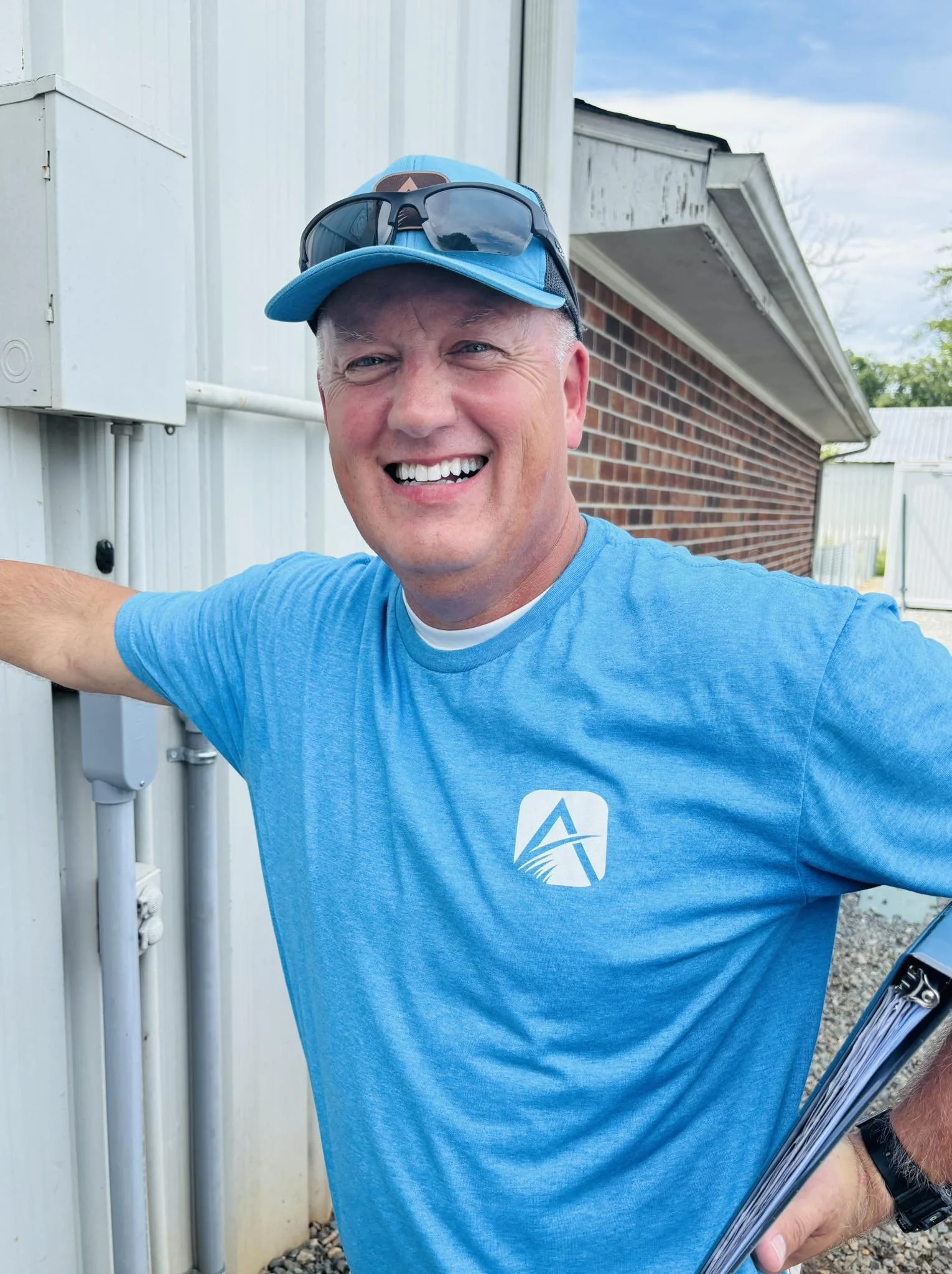 A smiling man wearing a light blue t-shirt, sunglasses on his blue cap, holding a clipboard, standing outside next to an electrical box.