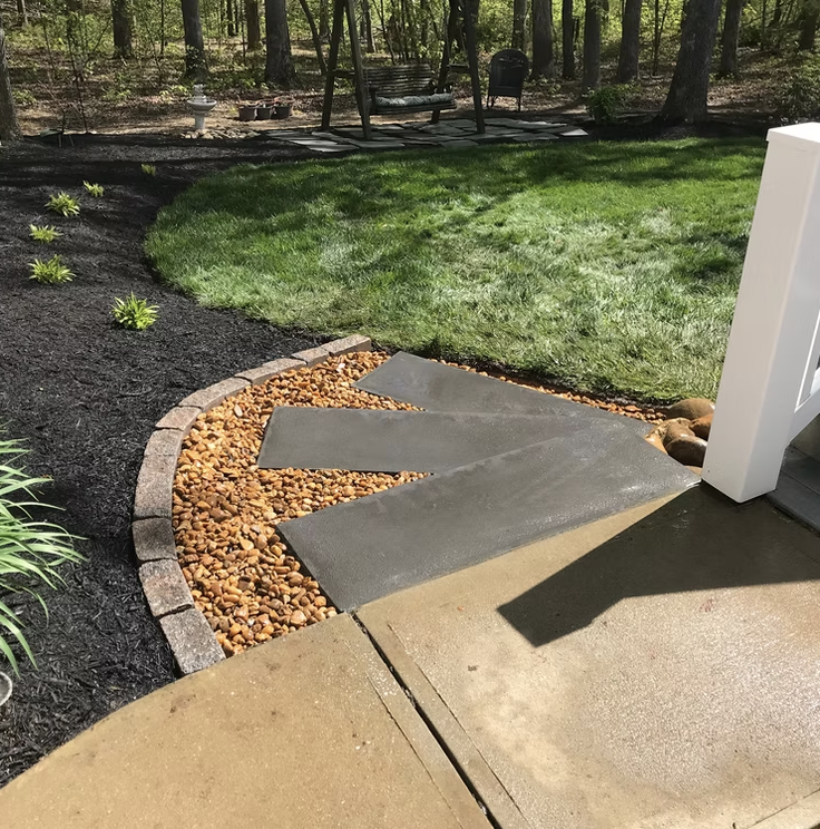 A landscaped backyard with a curved stone border, a small gravel area with stepping stones, green grass, and a wooded area in the background, with outdoor furniture and trees.
