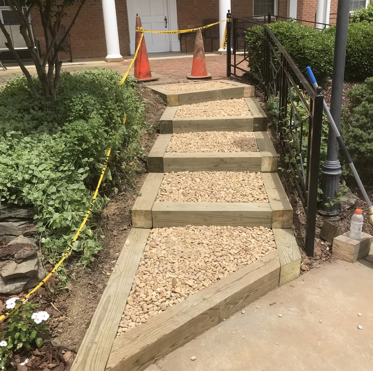 A new wooden and gravel pathway on a slope leading up to an entrance with steps, surrounded by greenery and orange traffic cones at the top.