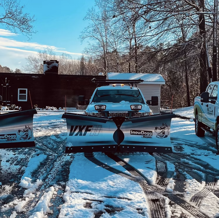 A snow removal vehicle with a snowplow attached to its front, parked on a snow-covered area with tire tracks. There is a truck on the right and a building in the background, with bare trees and a blue sky.